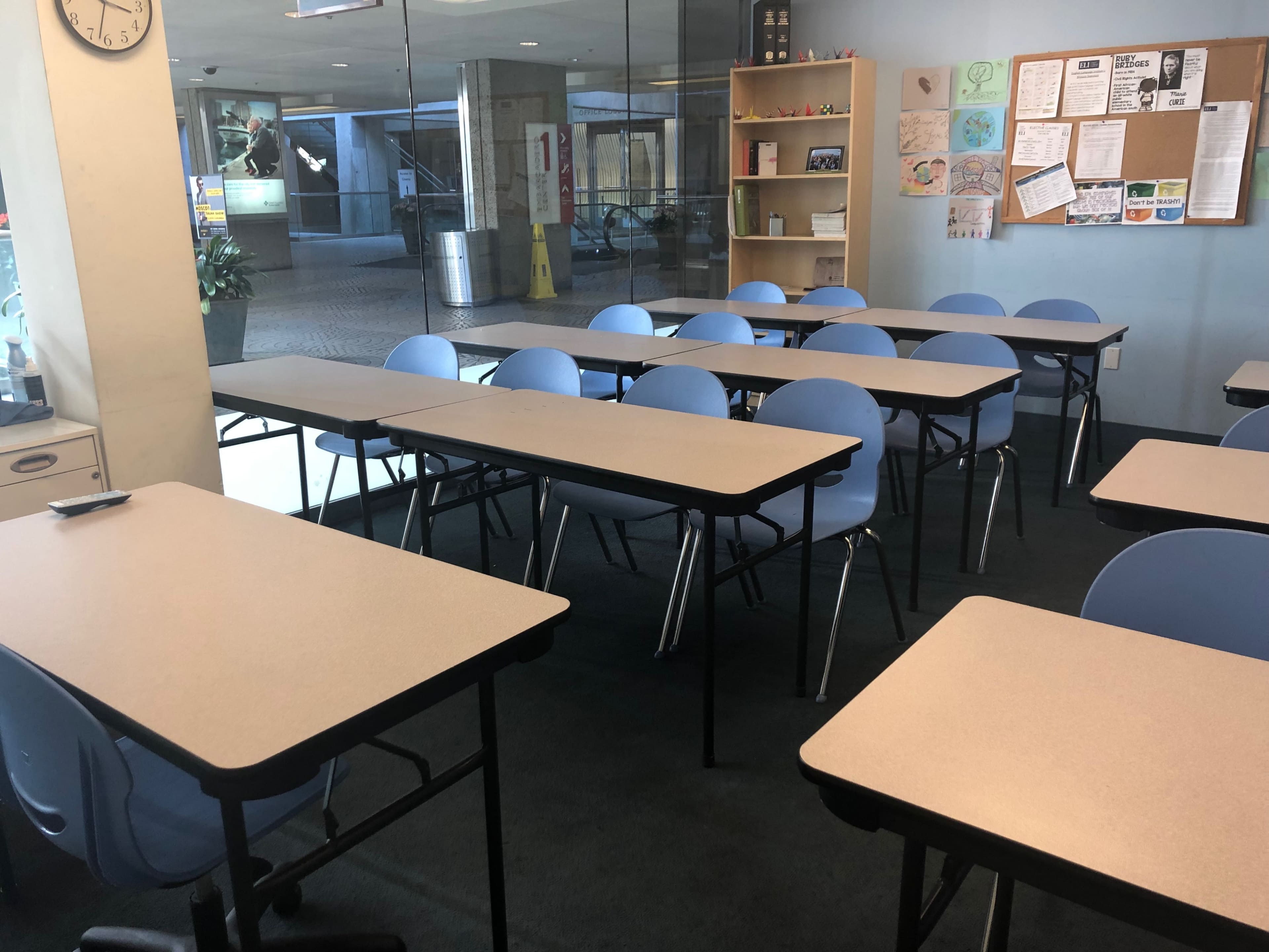 A classroom is set up with several rows of tables and blue chairs, illuminated by natural light coming through large windows.