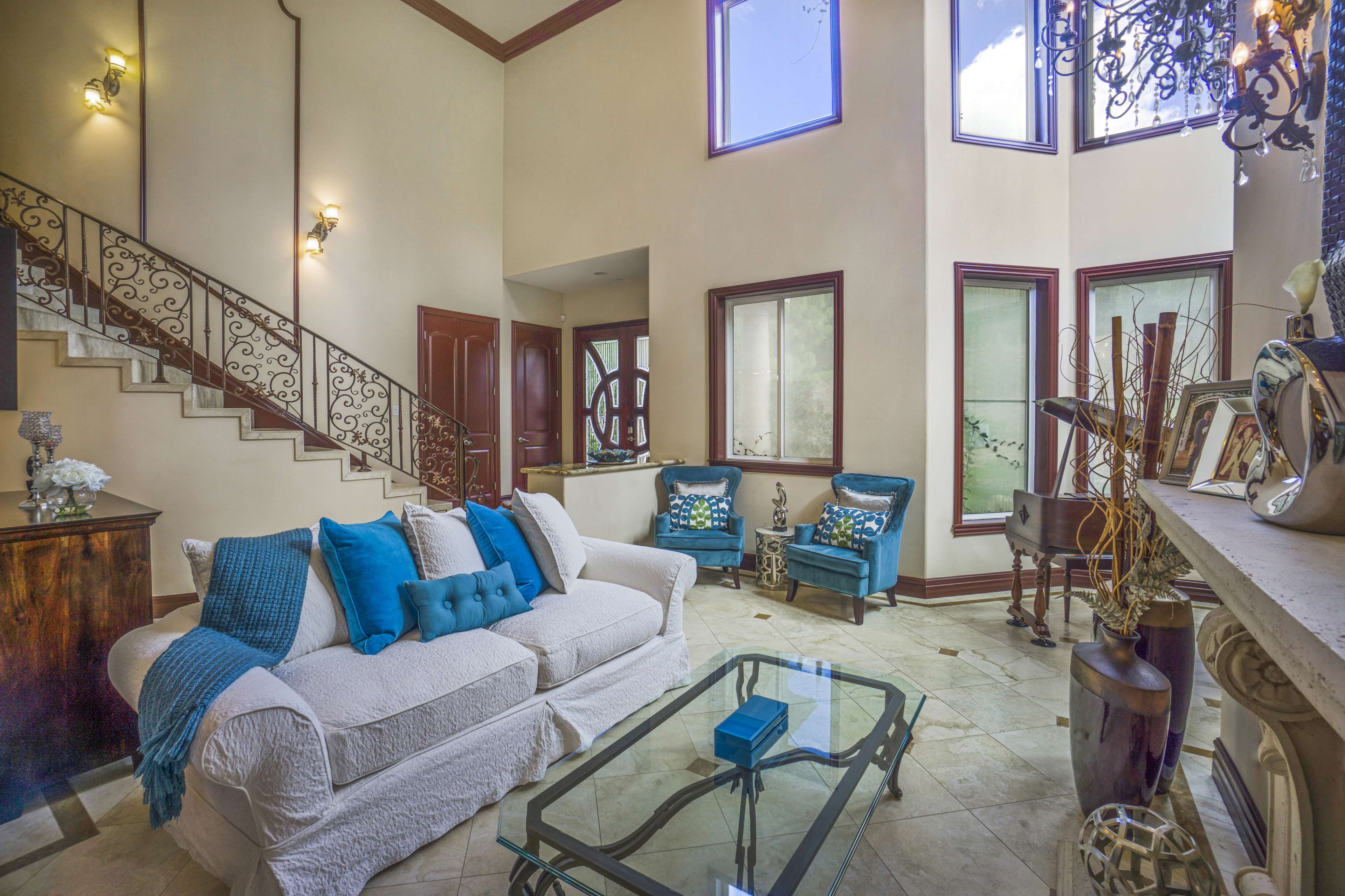 The image shows a well-lit living room featuring a white sofa with blue cushions, a glass coffee table, and large windows, along with a staircase and decorative elements.