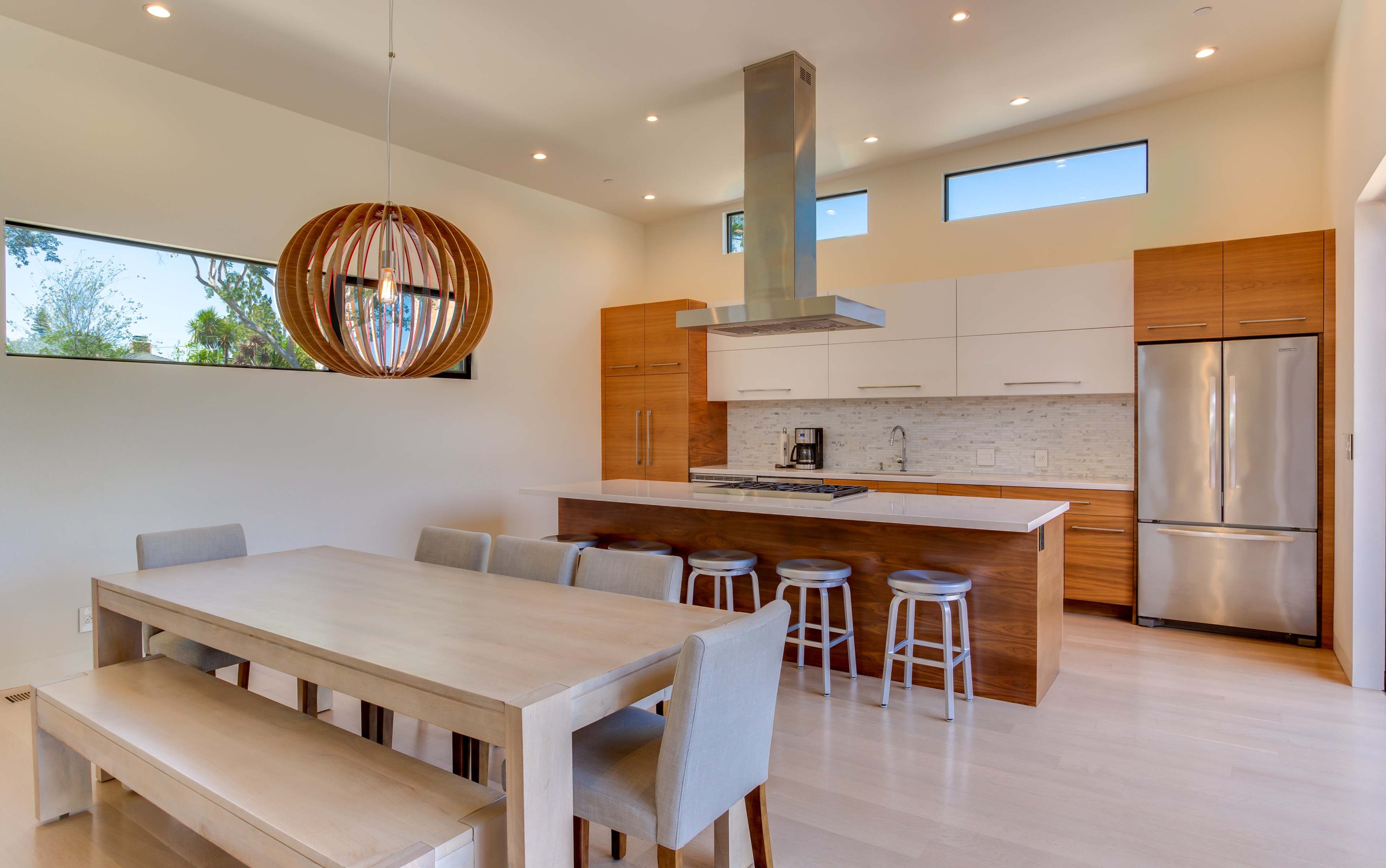 The image shows a modern kitchen and dining area featuring a central island with bar stools, a wooden dining table, and large windows for natural light.
