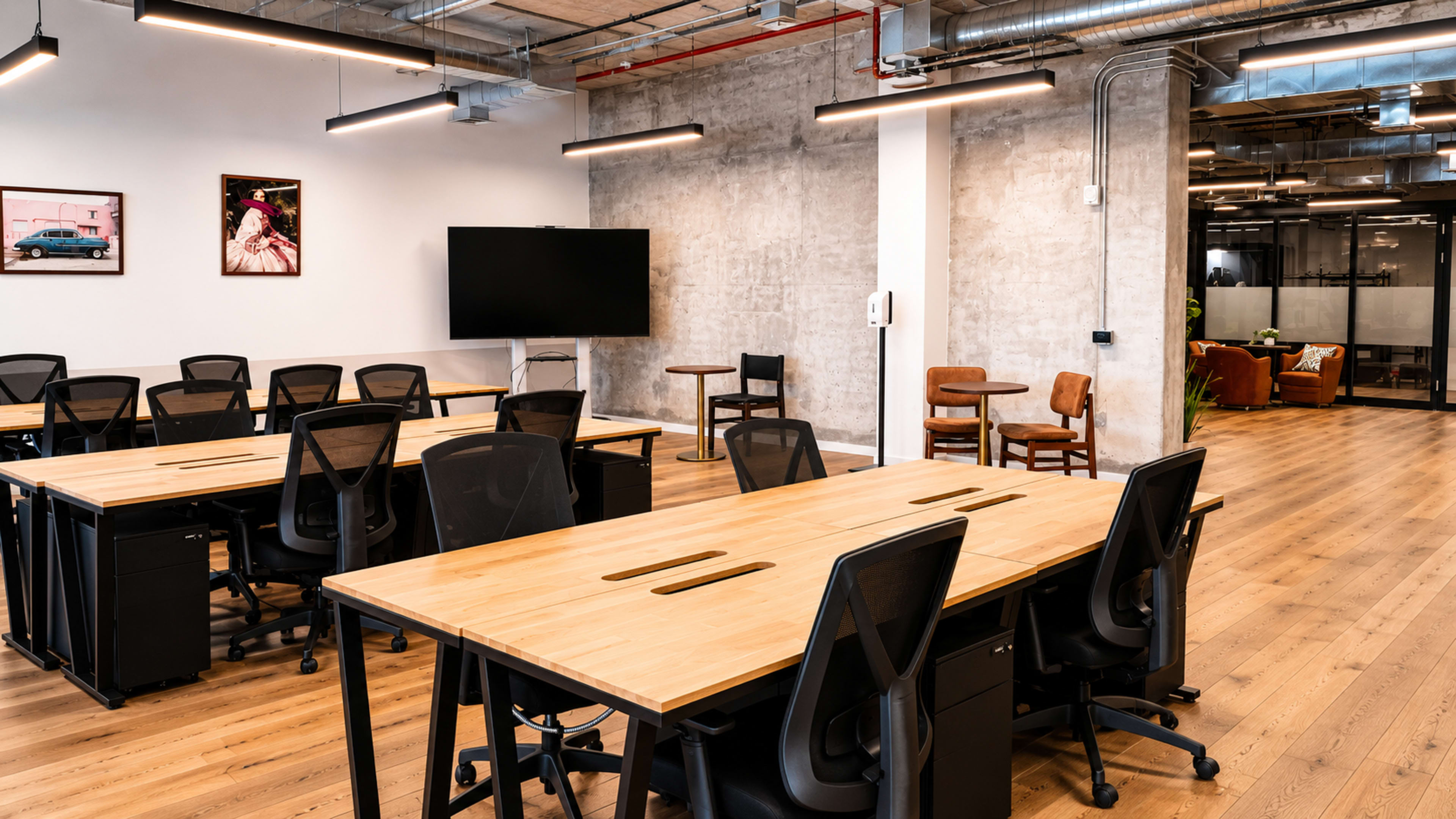 The image shows a modern office space with several wooden desks arranged in rows, a large wall-mounted screen, and a few seating areas in the background.