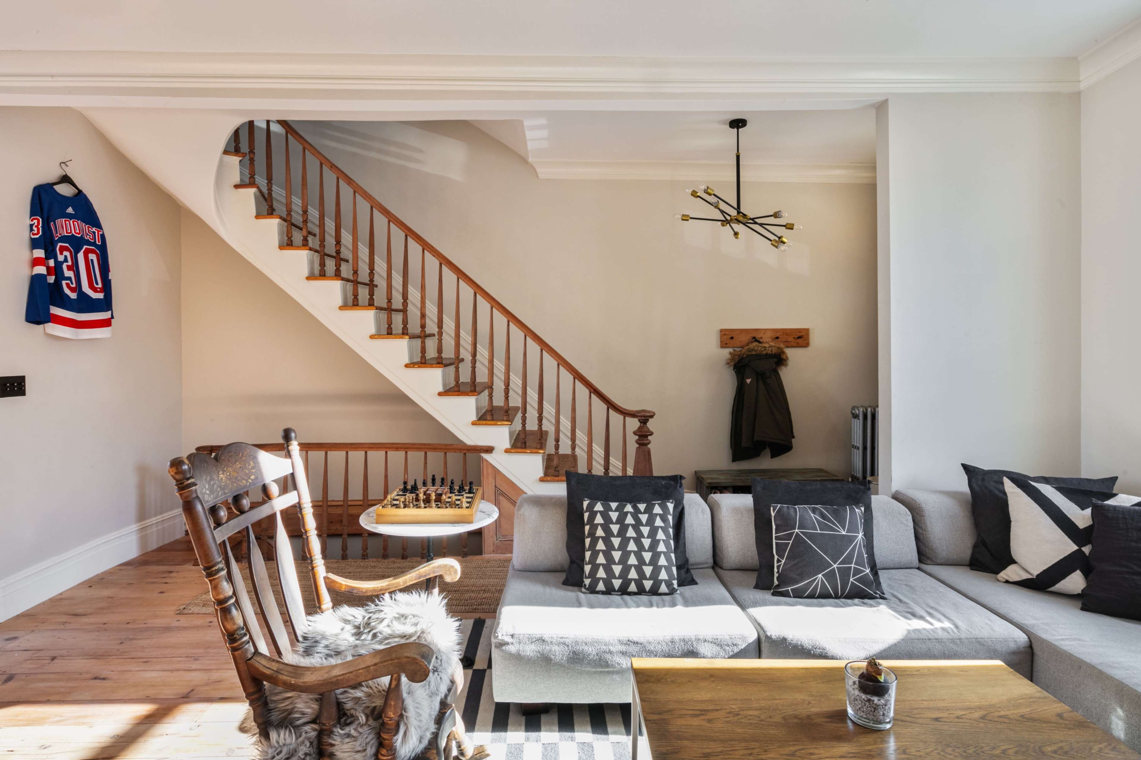 The image shows a living room with a gray sectional sofa, a wooden rocking chair, a staircase with a wooden railing, and a hockey jersey hanging on the wall.