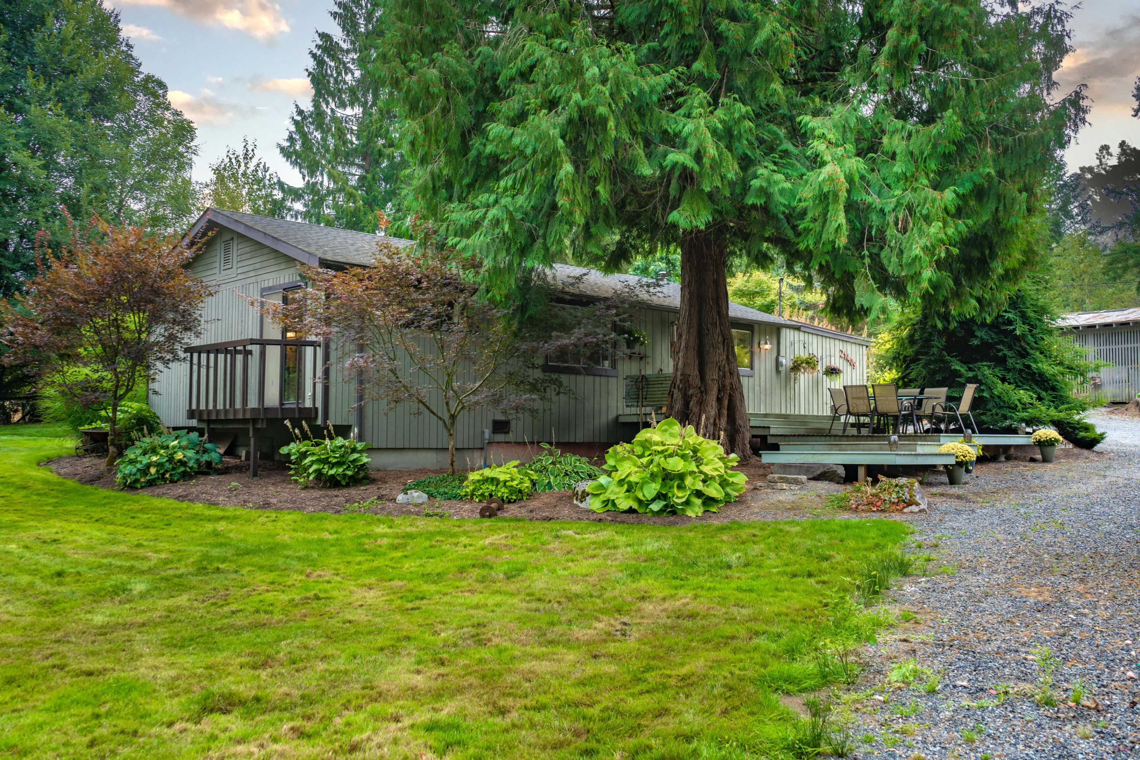 A gray single-story house with a deck and surrounding greenery is situated on a landscaped property.