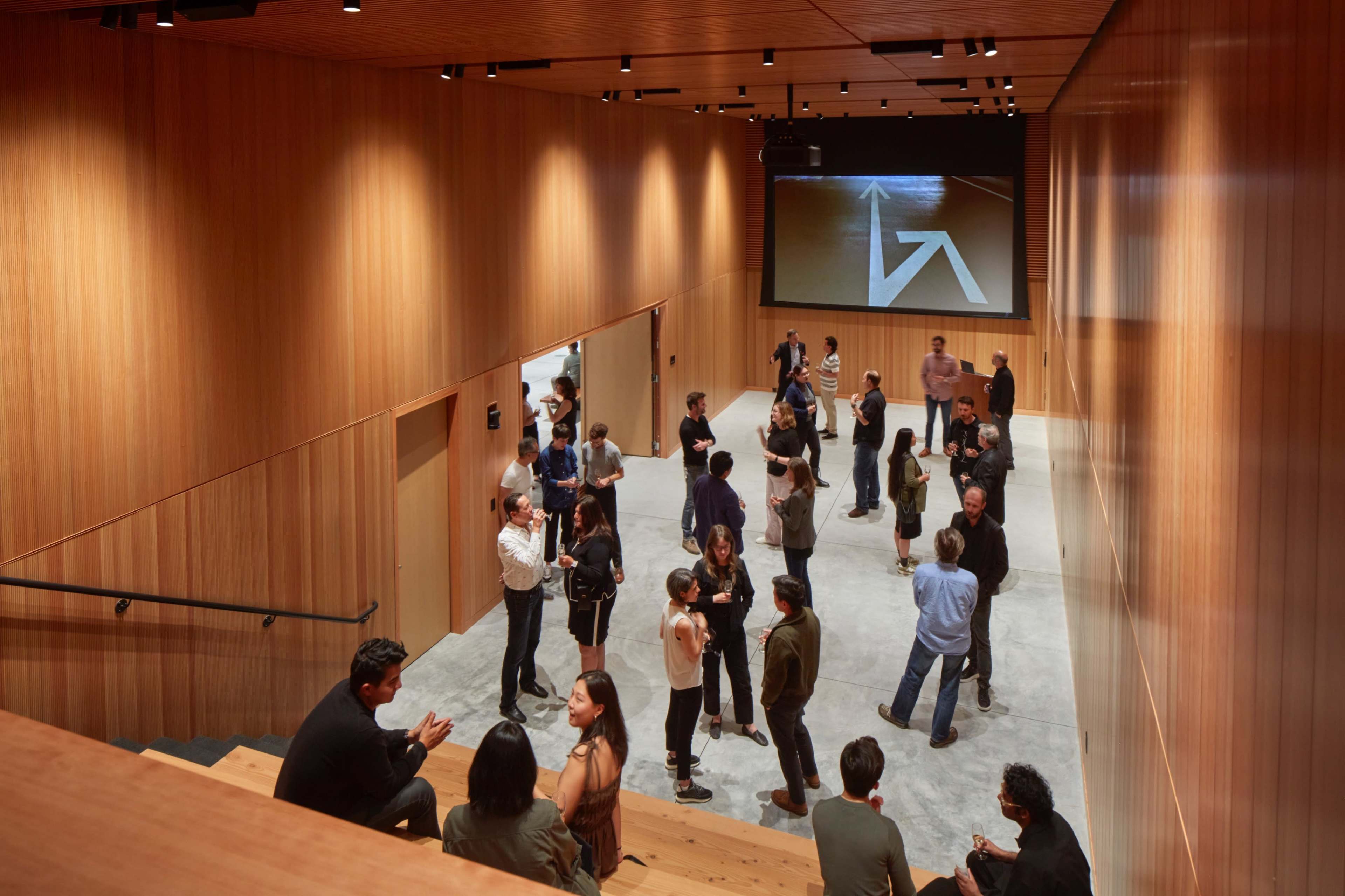 A group of people is gathered in a modern wooden interior space with a large screen displaying an arrow.