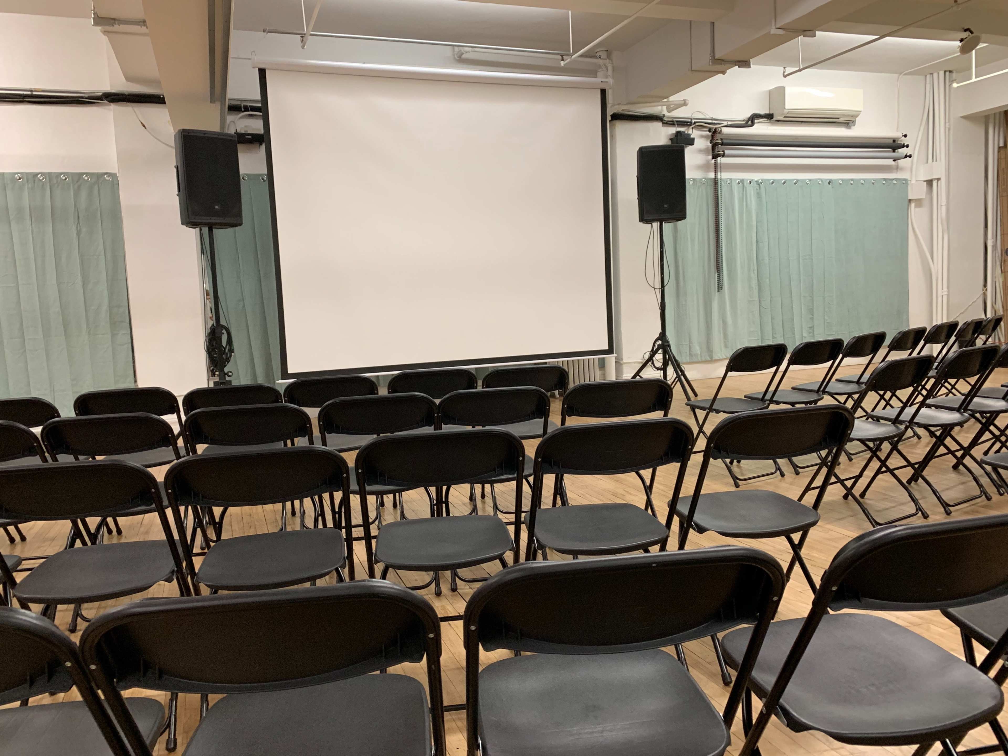 A row of black folding chairs facing a blank screen in a room with light green curtains.