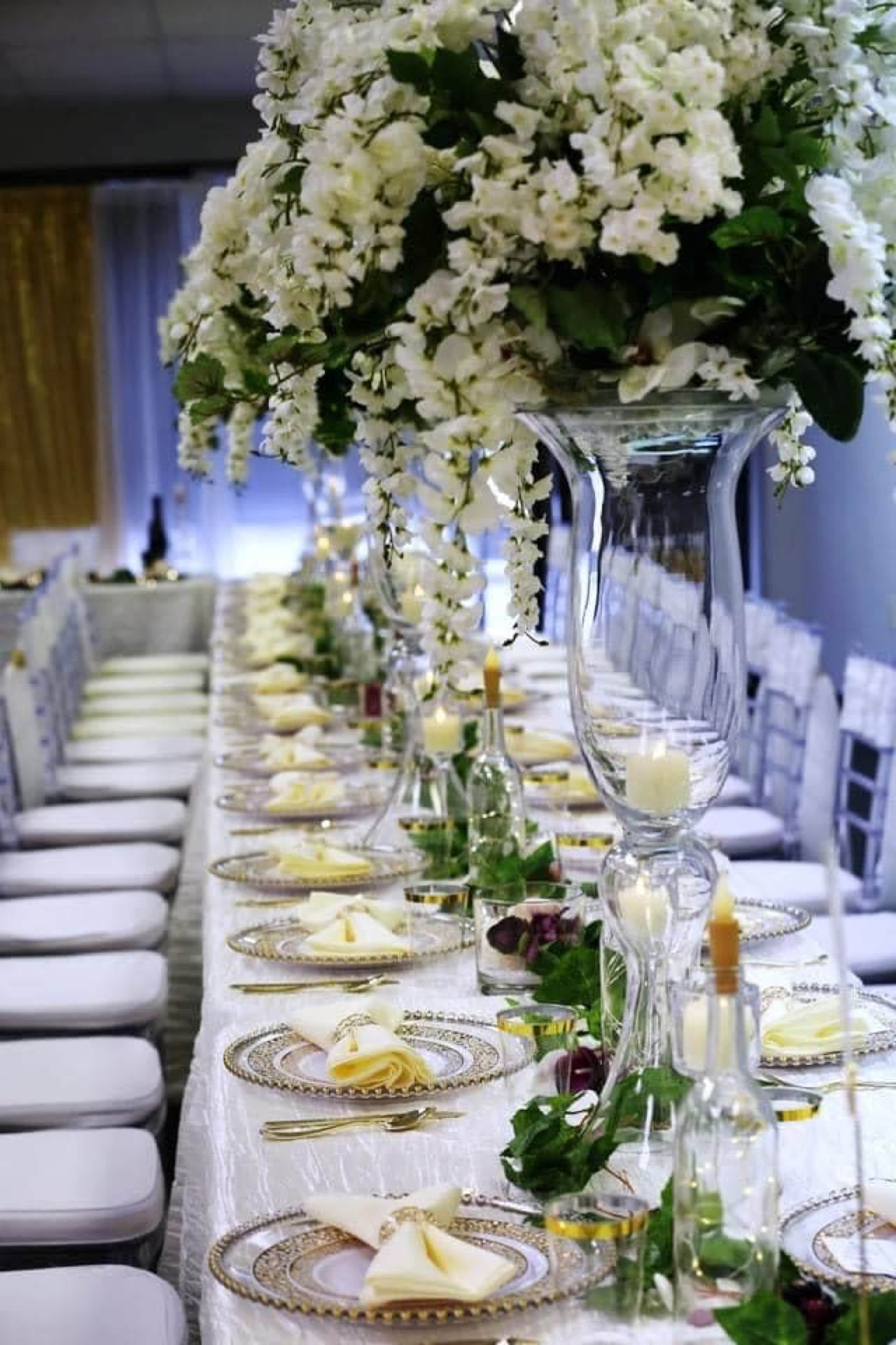 The image shows a long dining table elegantly set with white chairs, featuring decorative floral arrangements and candles in tall glass vases.
