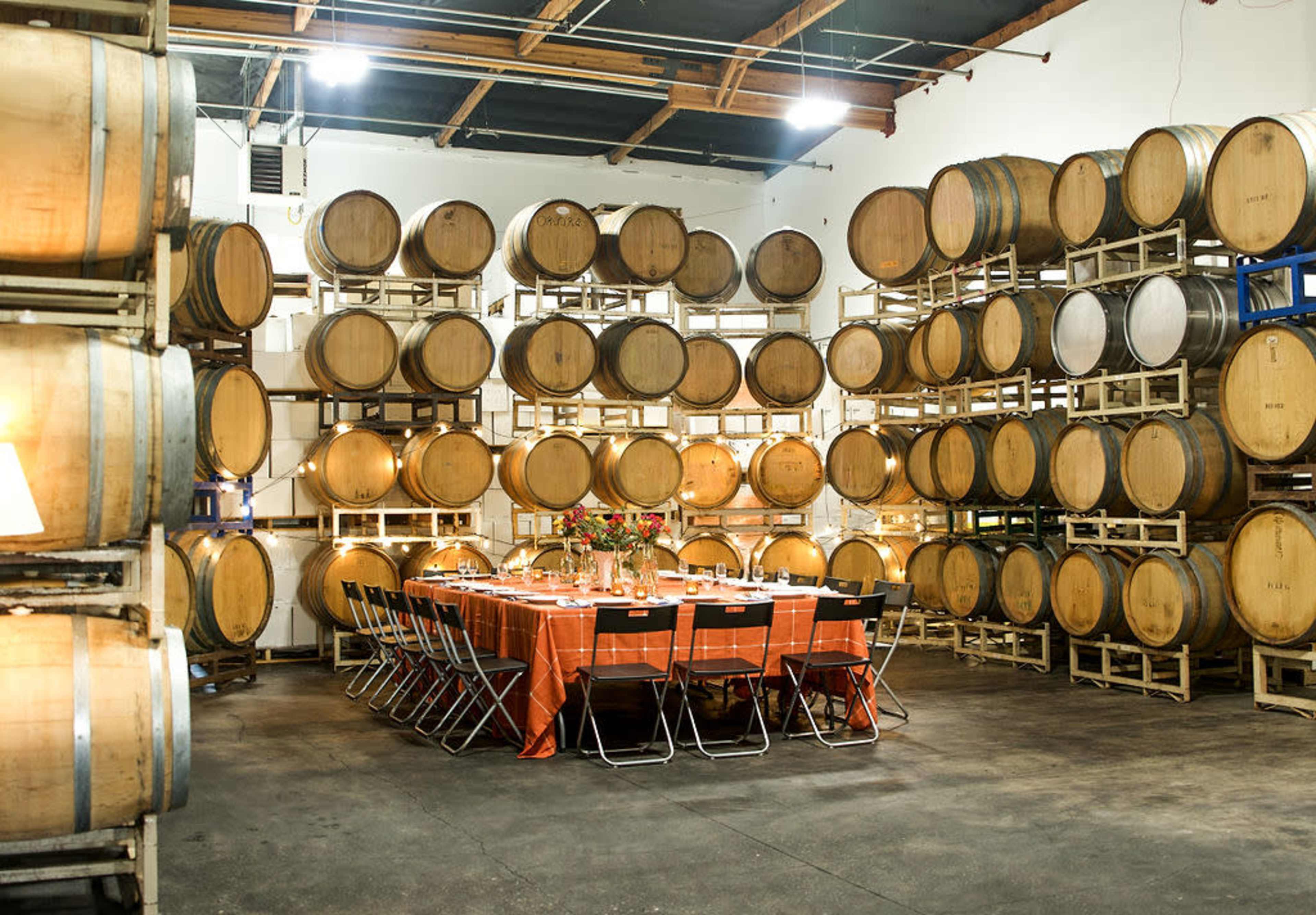 A long table is set for an event surrounded by rows of wooden wine barrels in a warehouse.