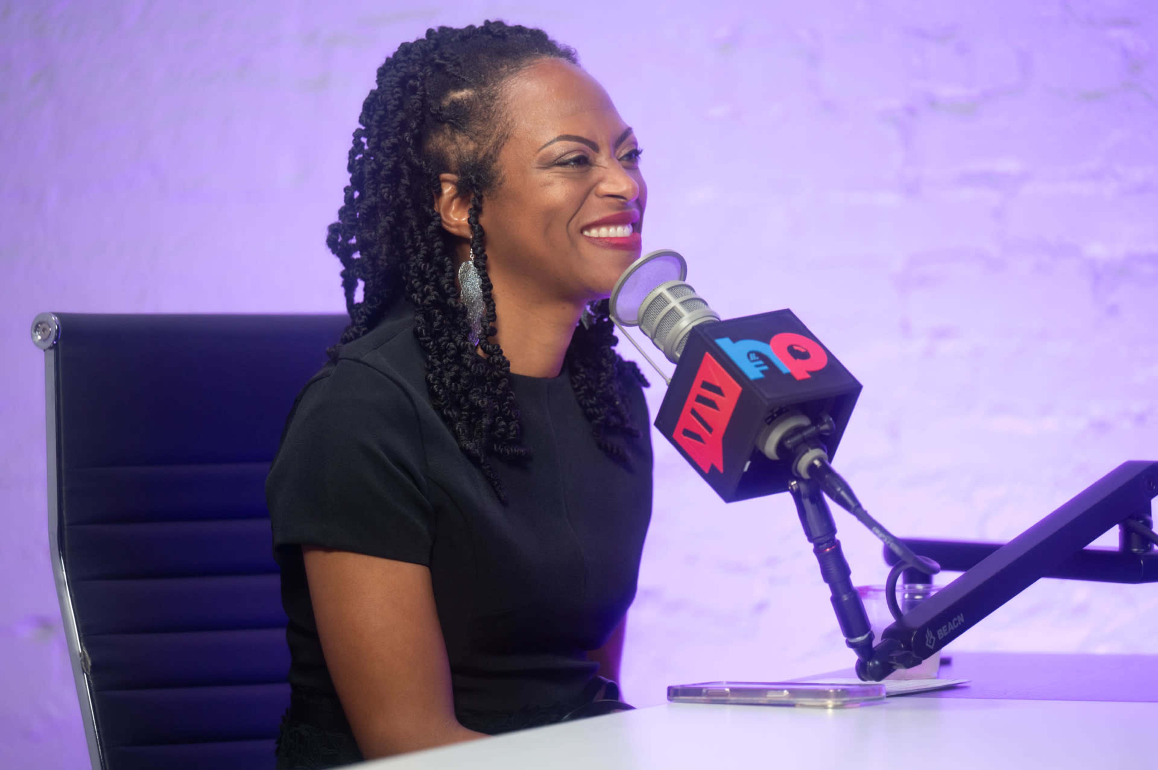 A woman sits at a microphone in a studio with a purple background, smiling while speaking.