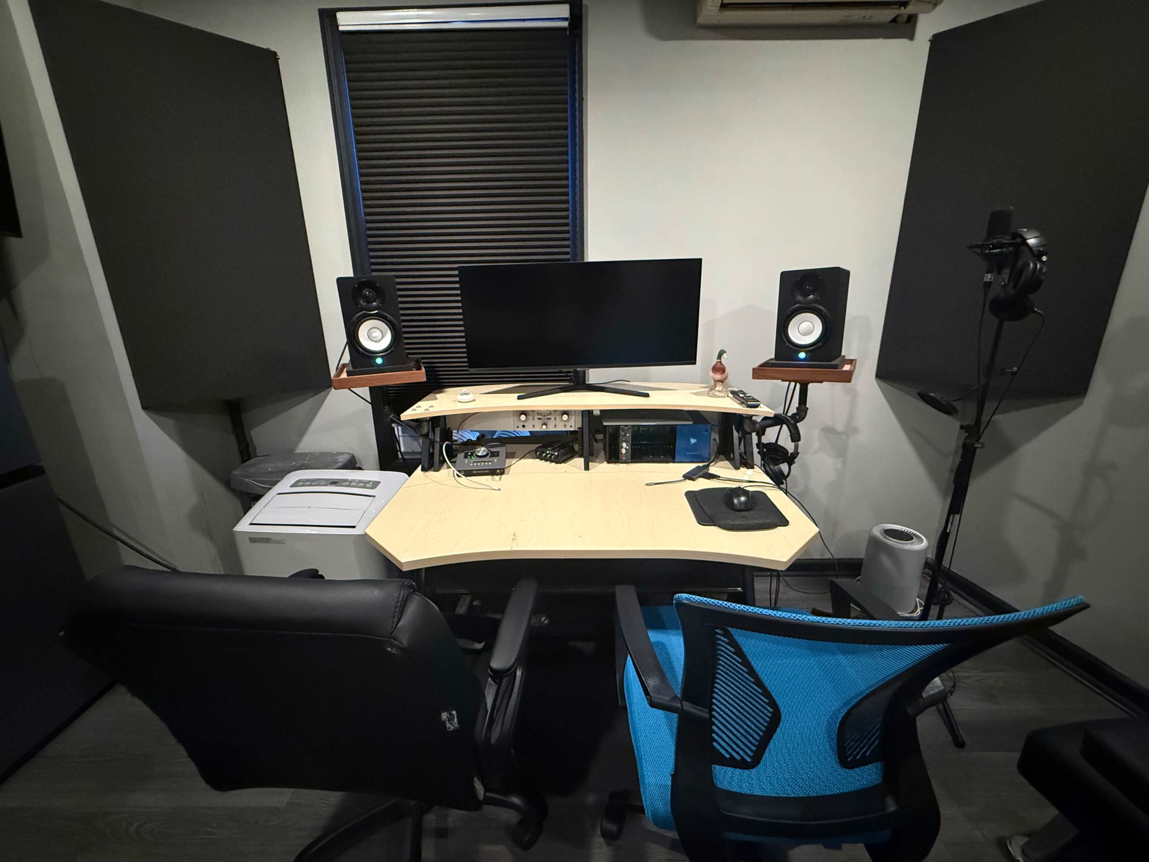 The image shows a recording studio setup featuring a desk with a computer monitor, two speakers, a microphone on a stand, and two office chairs.