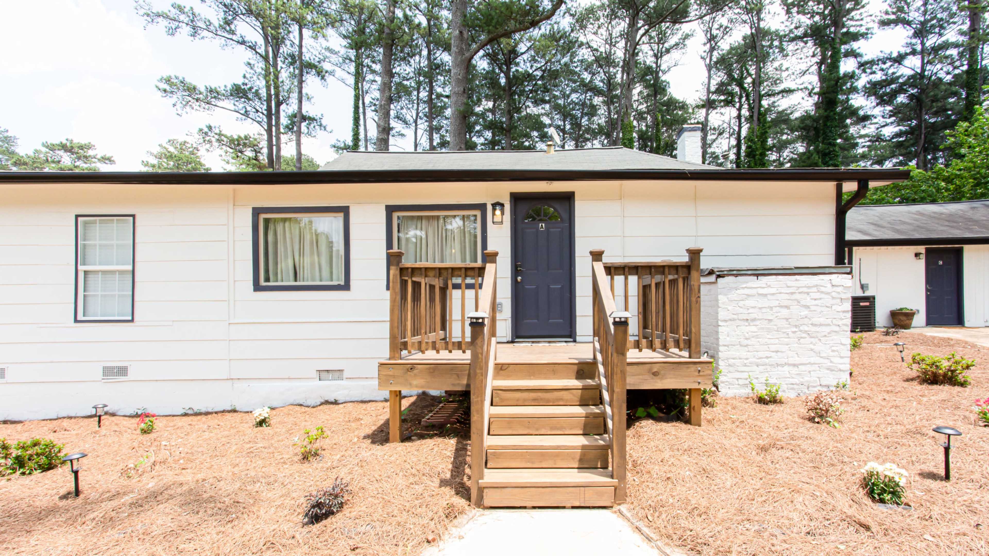 A single-story home with a wooden front porch and steps, surrounded by mulch and small plants.