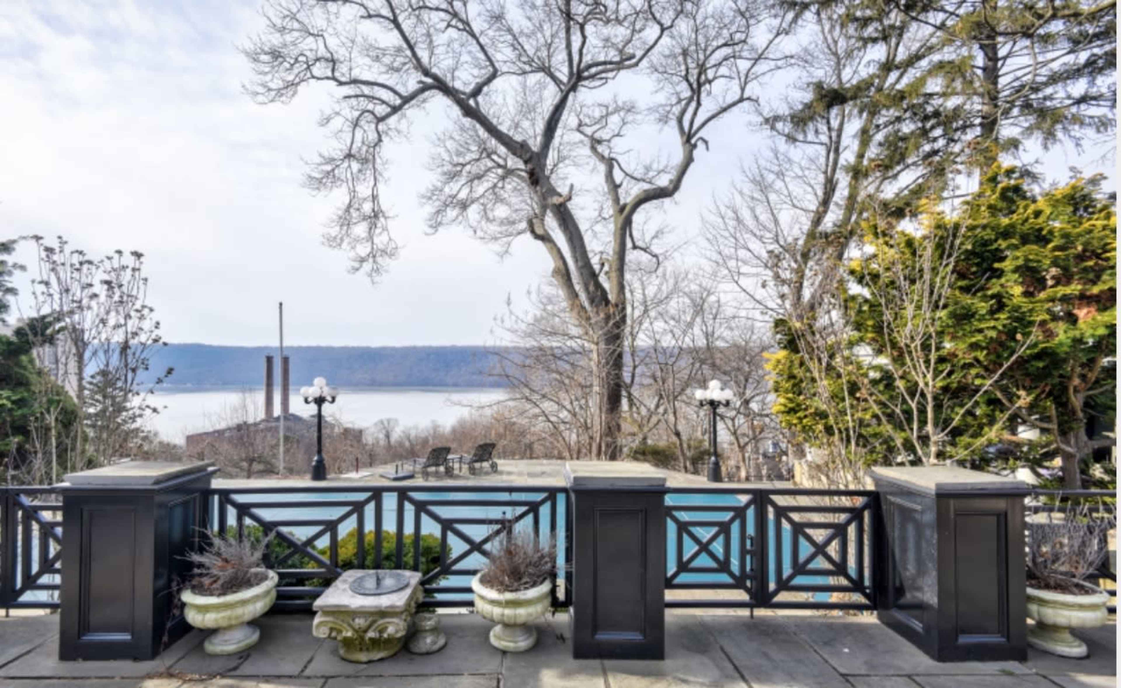 The image shows a view of a pool area surrounded by planters and trees, overlooking a river with a cloudy sky above.