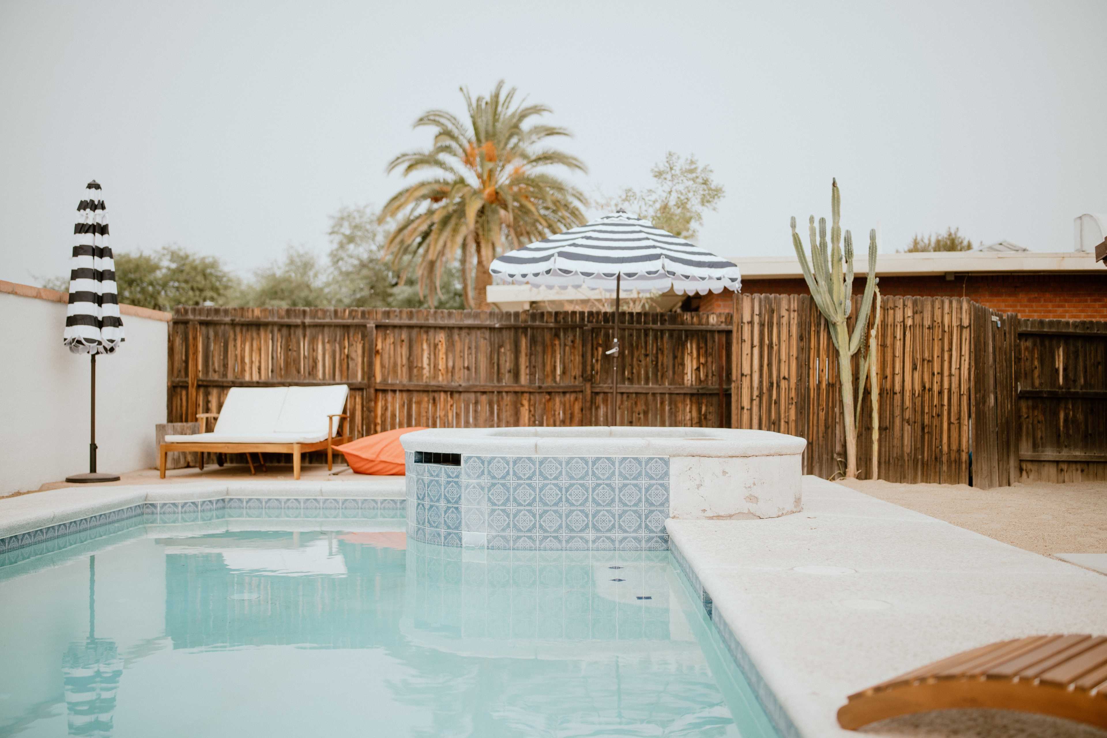 A swimming pool surrounded by a wooden fence, with lounge chairs, a striped umbrella, and a cactus in the background.