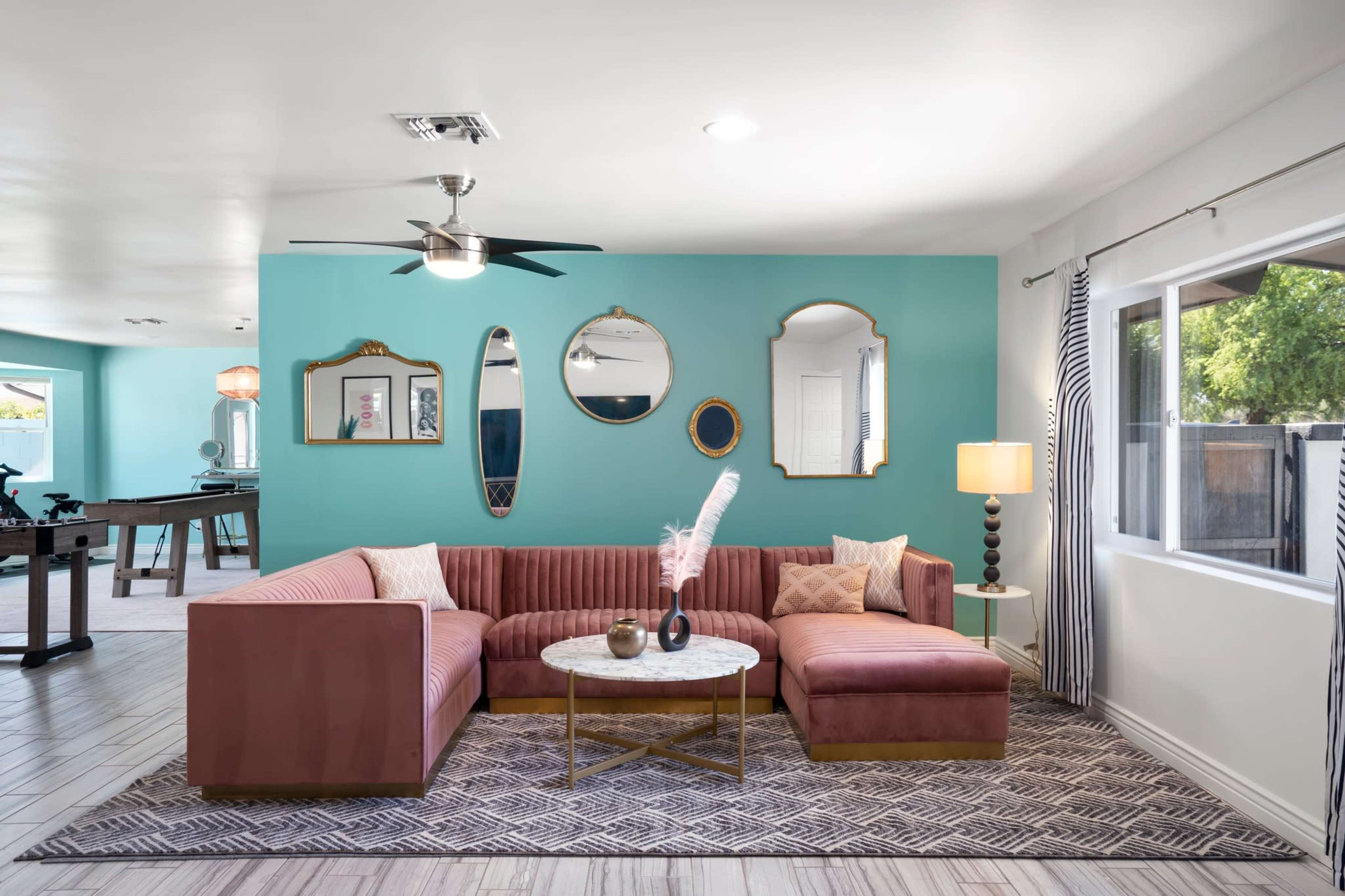 A modern living room with a large pink sectional couch, a round coffee table, and turquoise walls, featuring decorative mirrors and a lamp.