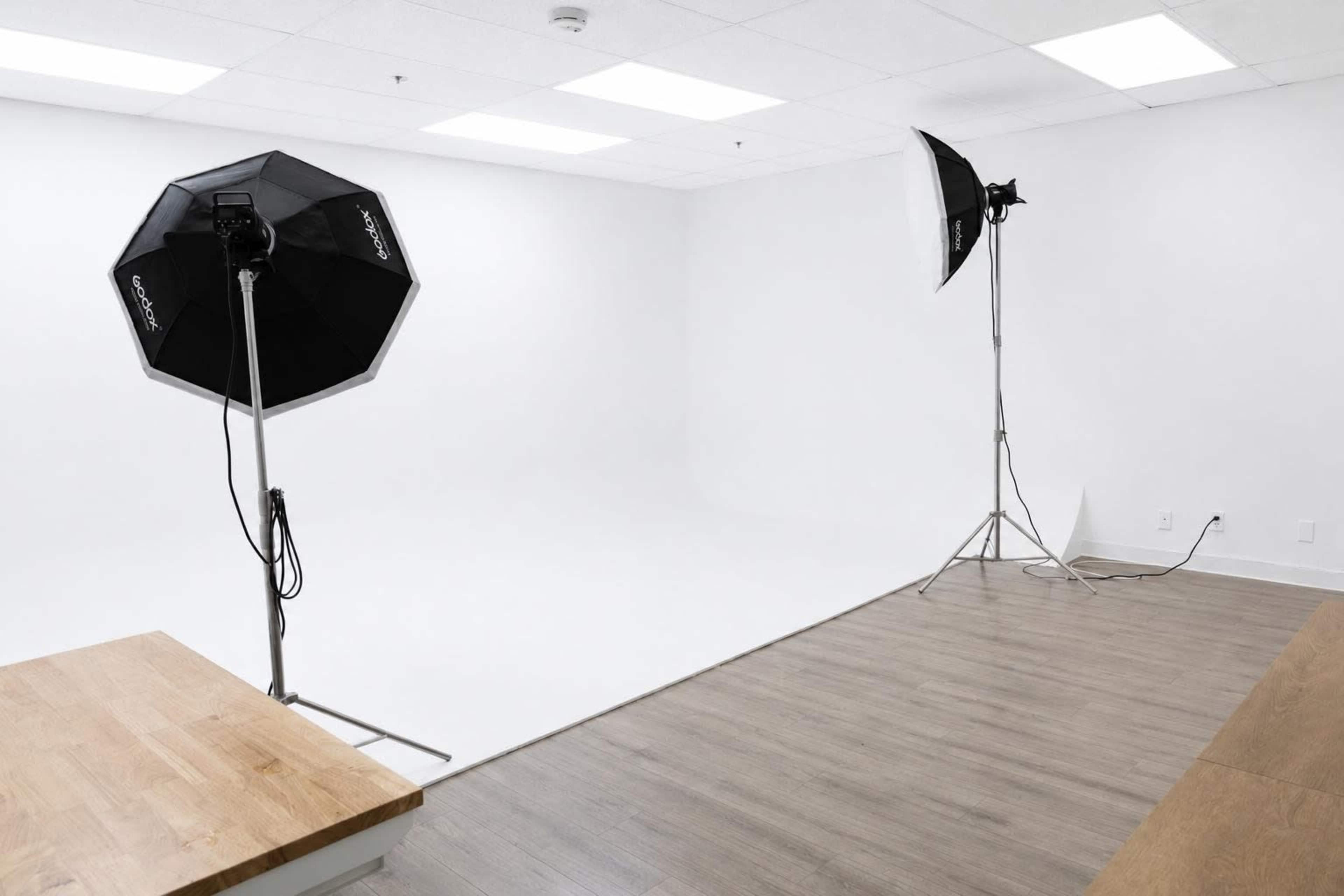 The image shows a spacious photography studio with two lighting setups on stands, a seamless backdrop, and wooden flooring.