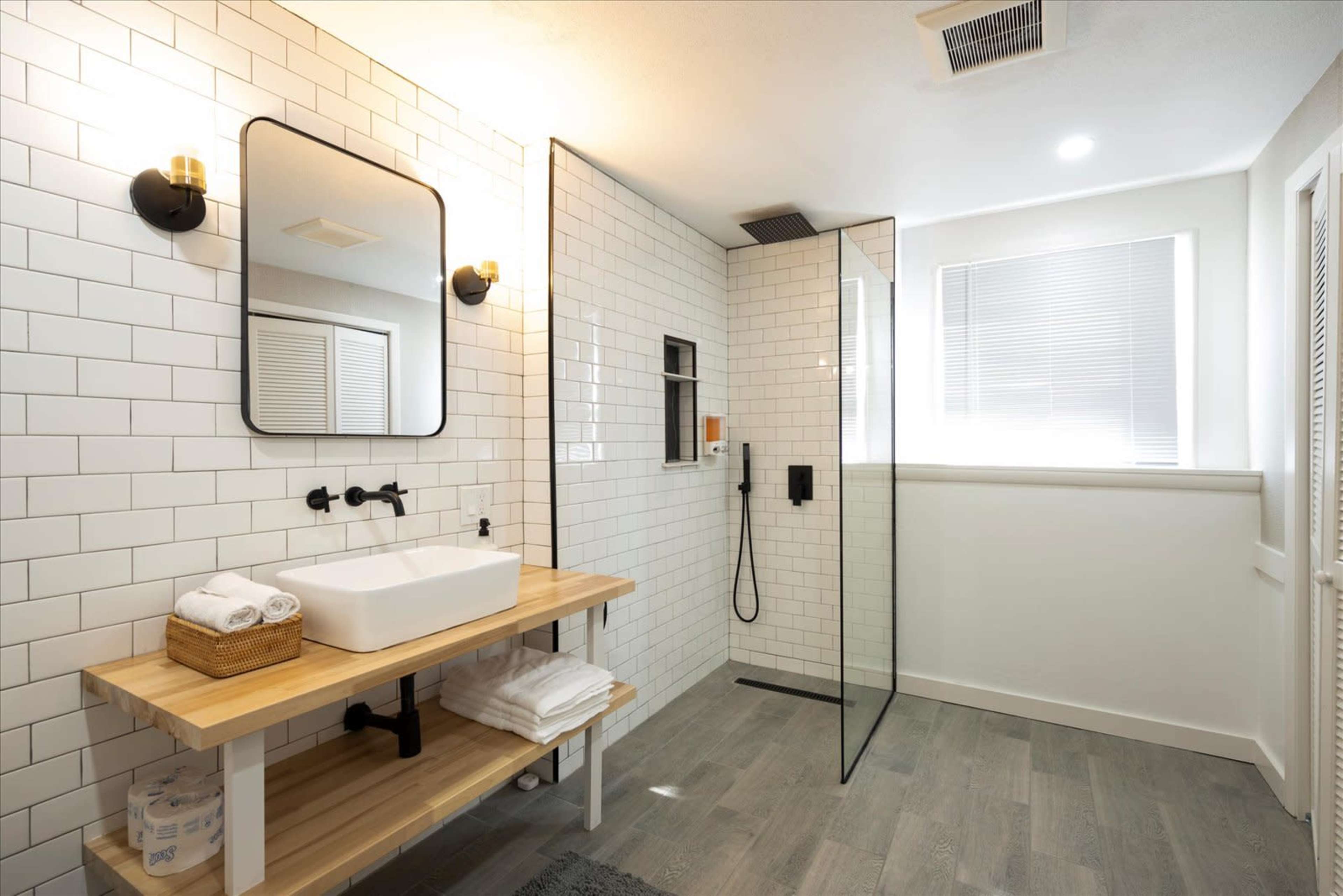 The image shows a modern bathroom featuring a walk-in shower, a wall-mounted mirror, a wooden countertop with a sink, and neatly arranged towels.