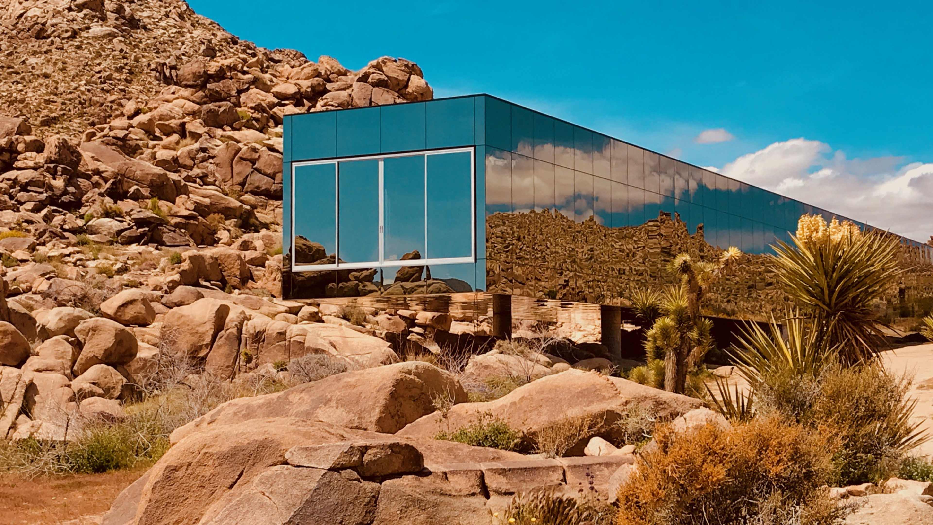 A modern glass building reflects the surrounding rocky landscape and desert vegetation under a blue sky.