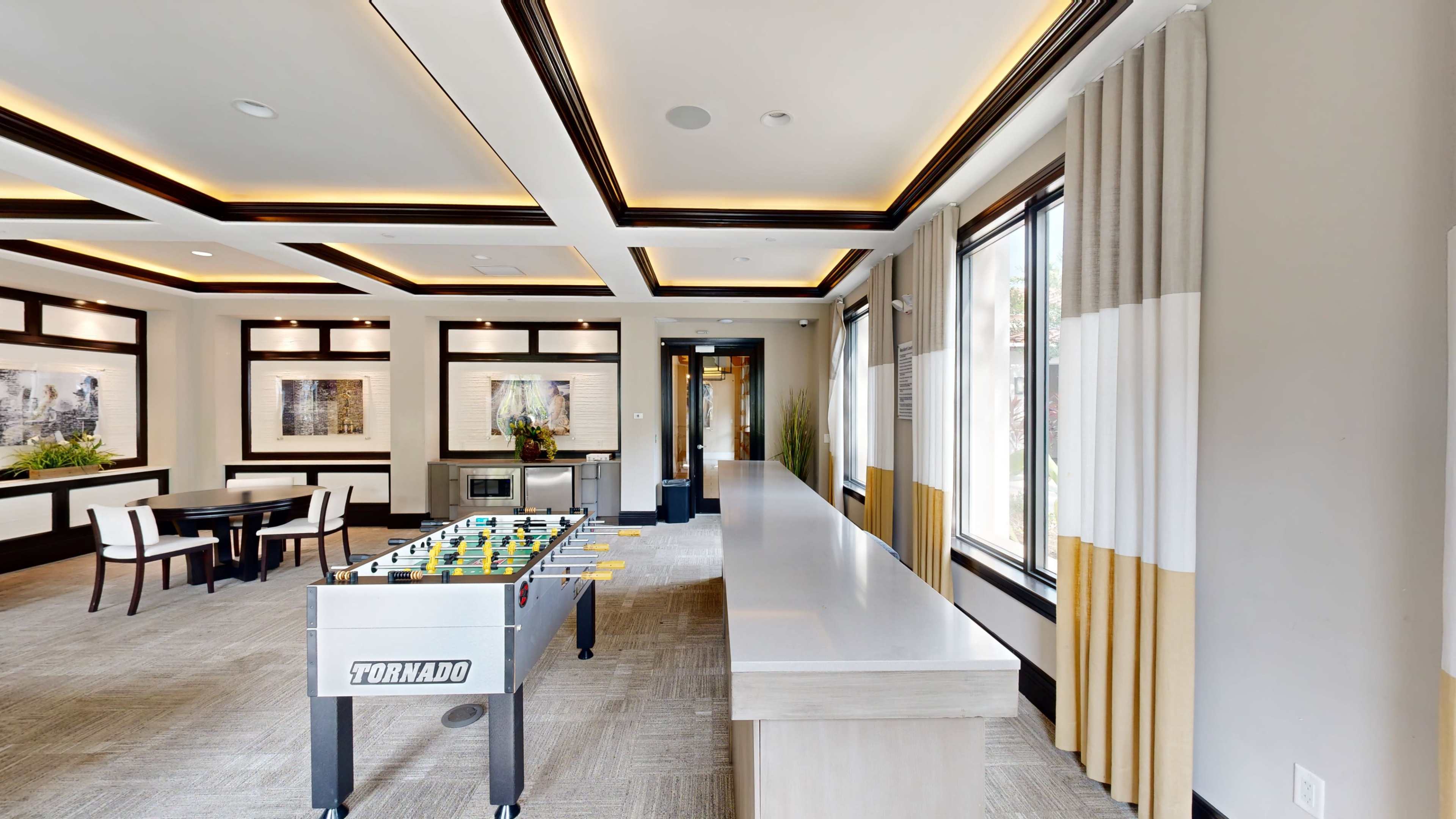 The image shows a modern game room featuring a foosball table, a long countertop, and large windows with decorative curtains.