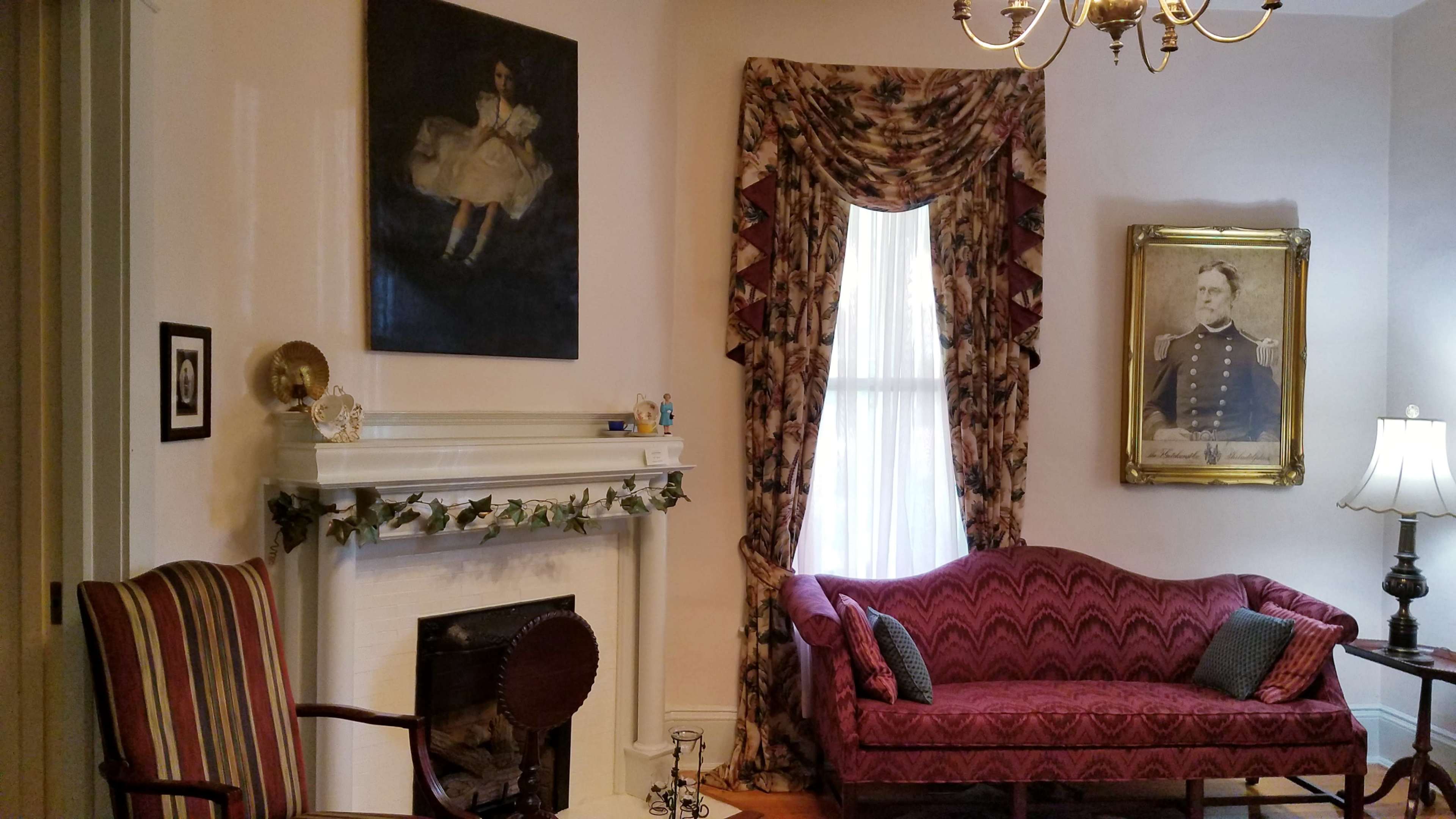 The image shows a living room featuring a purple upholstered sofa, a white fireplace decorated with garland, and a large window adorned with patterned curtains.