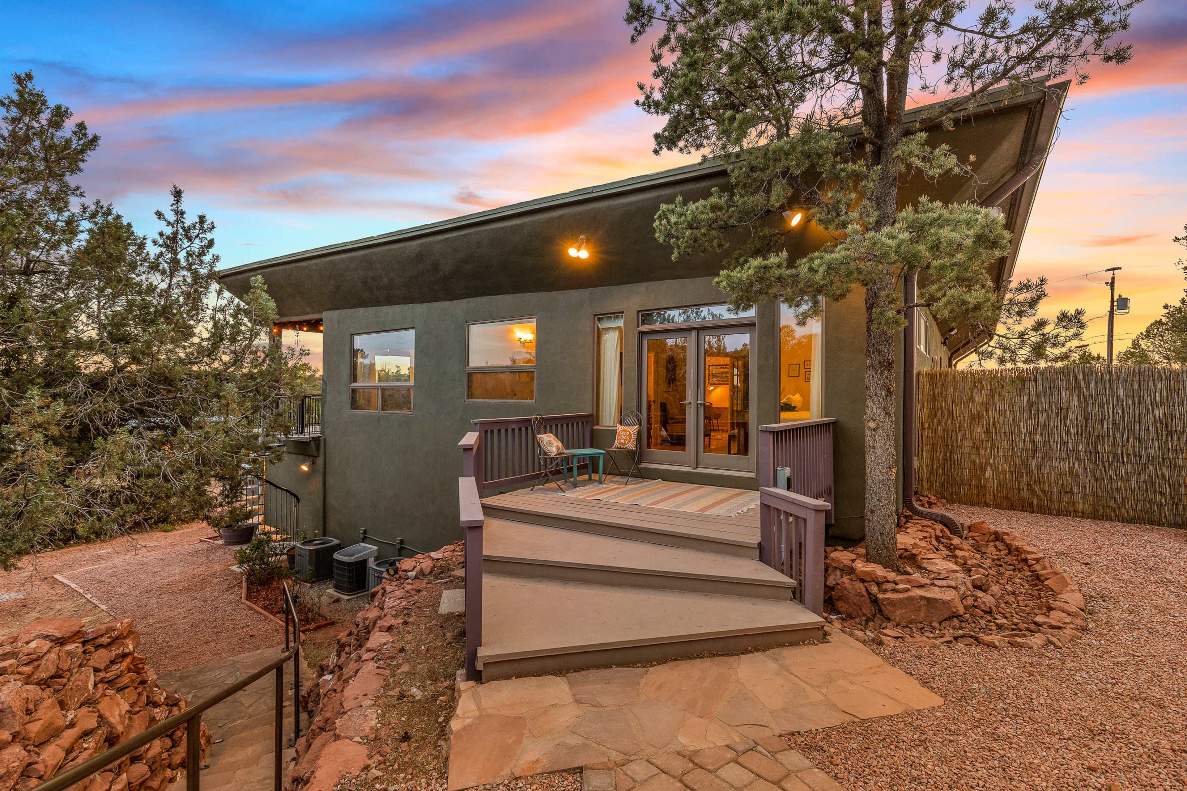 A modern house with large windows and a deck sits among trees at sunset, reflecting vibrant colors in the sky.