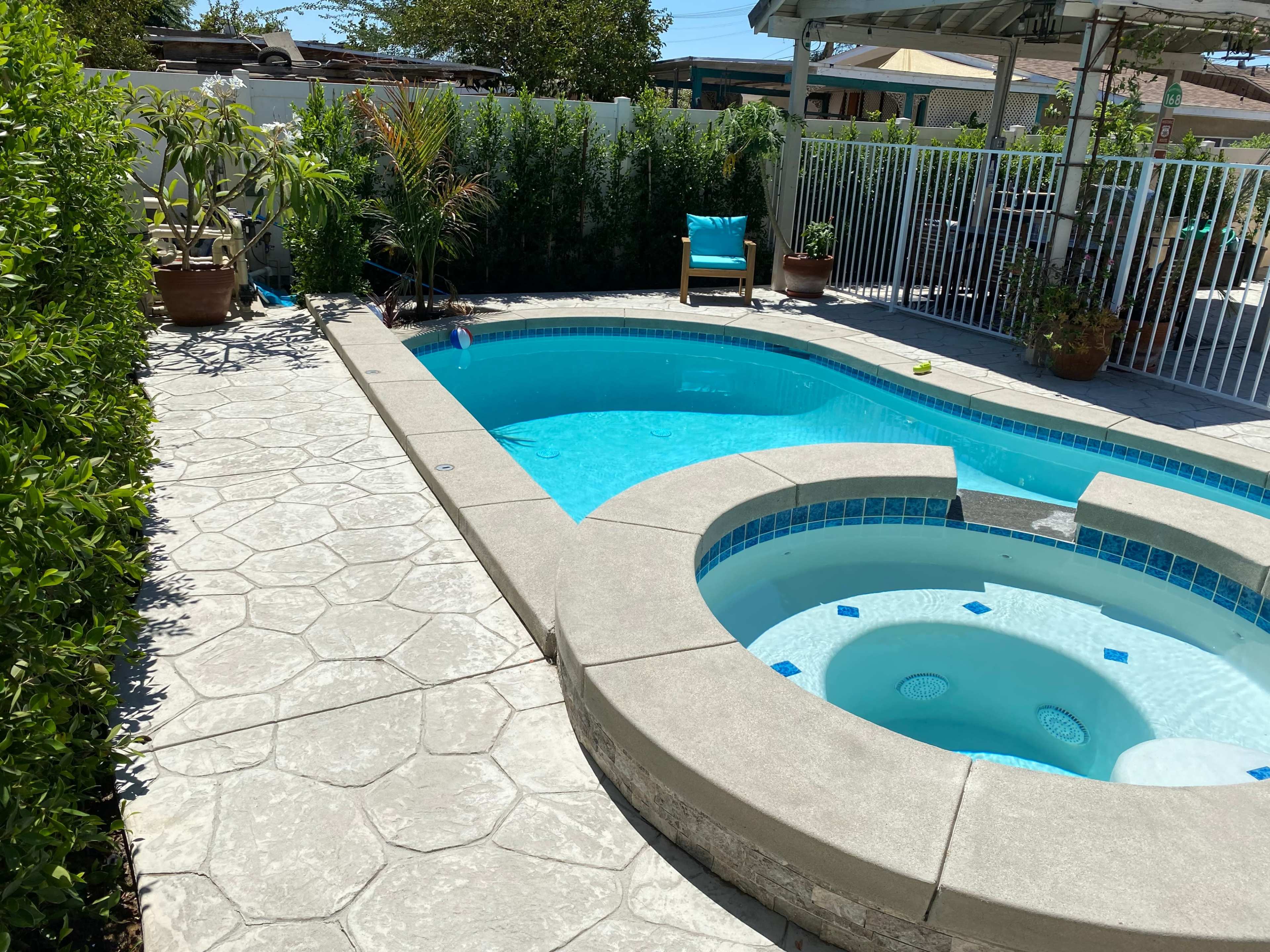 The image shows a swimming pool and a spa area, surrounded by a landscaped outdoor space with greenery and patio furniture.