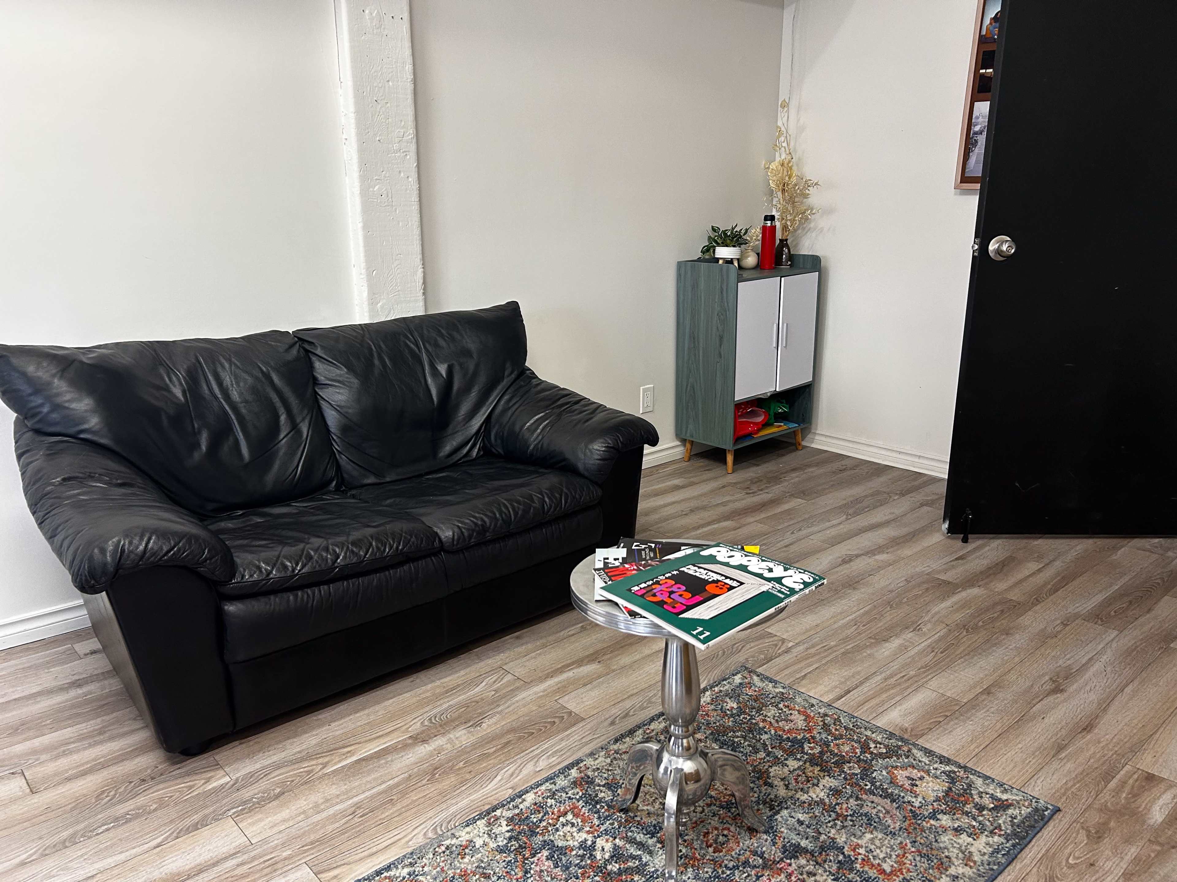The image shows a small living area with a black leather couch, a round table, and a cabinet against the wall.
