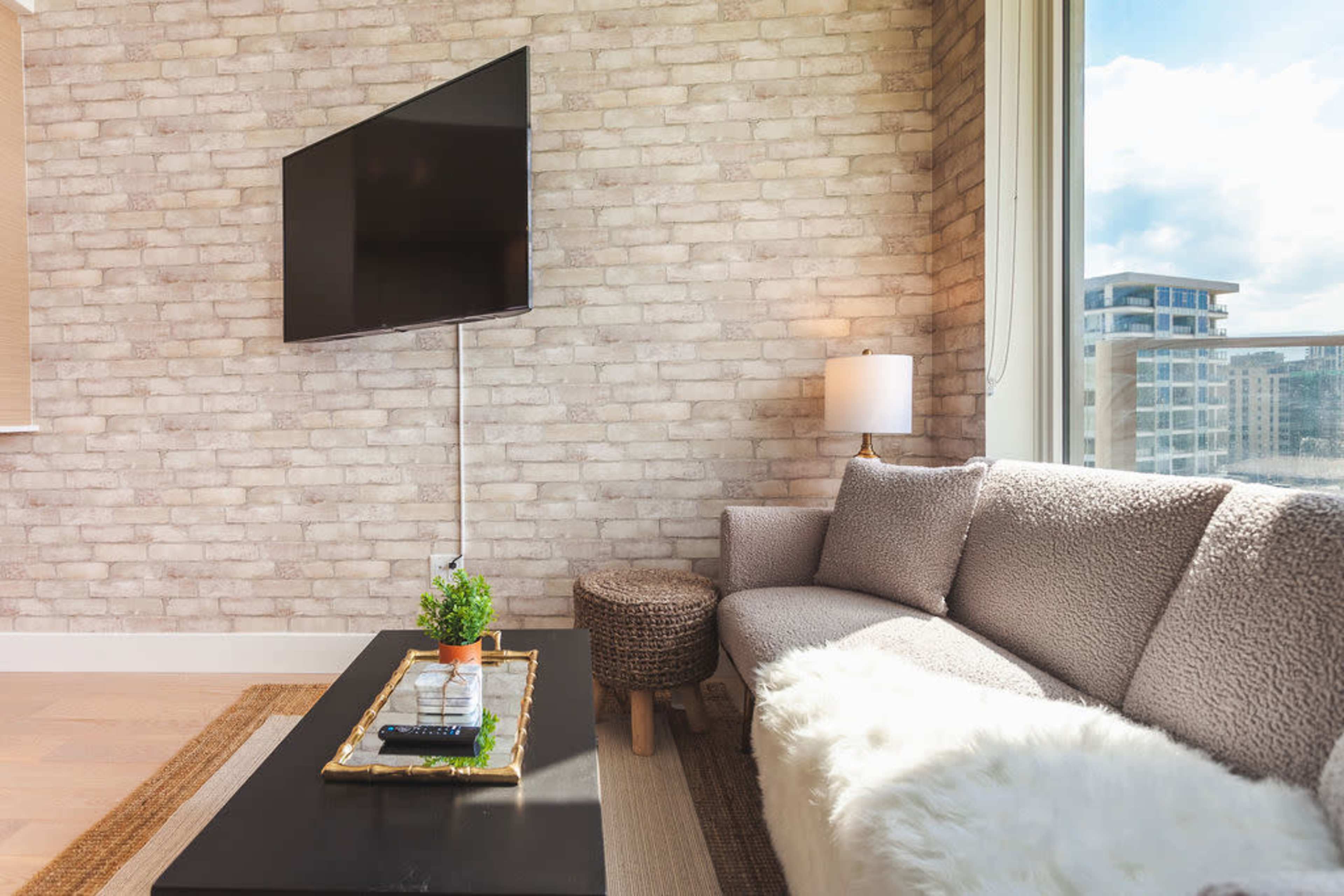A modern living room features a textured brick wall, a flat-screen TV mounted above a minimalist coffee table, and a cozy seating area with a light-colored sofa and a decorative plant.