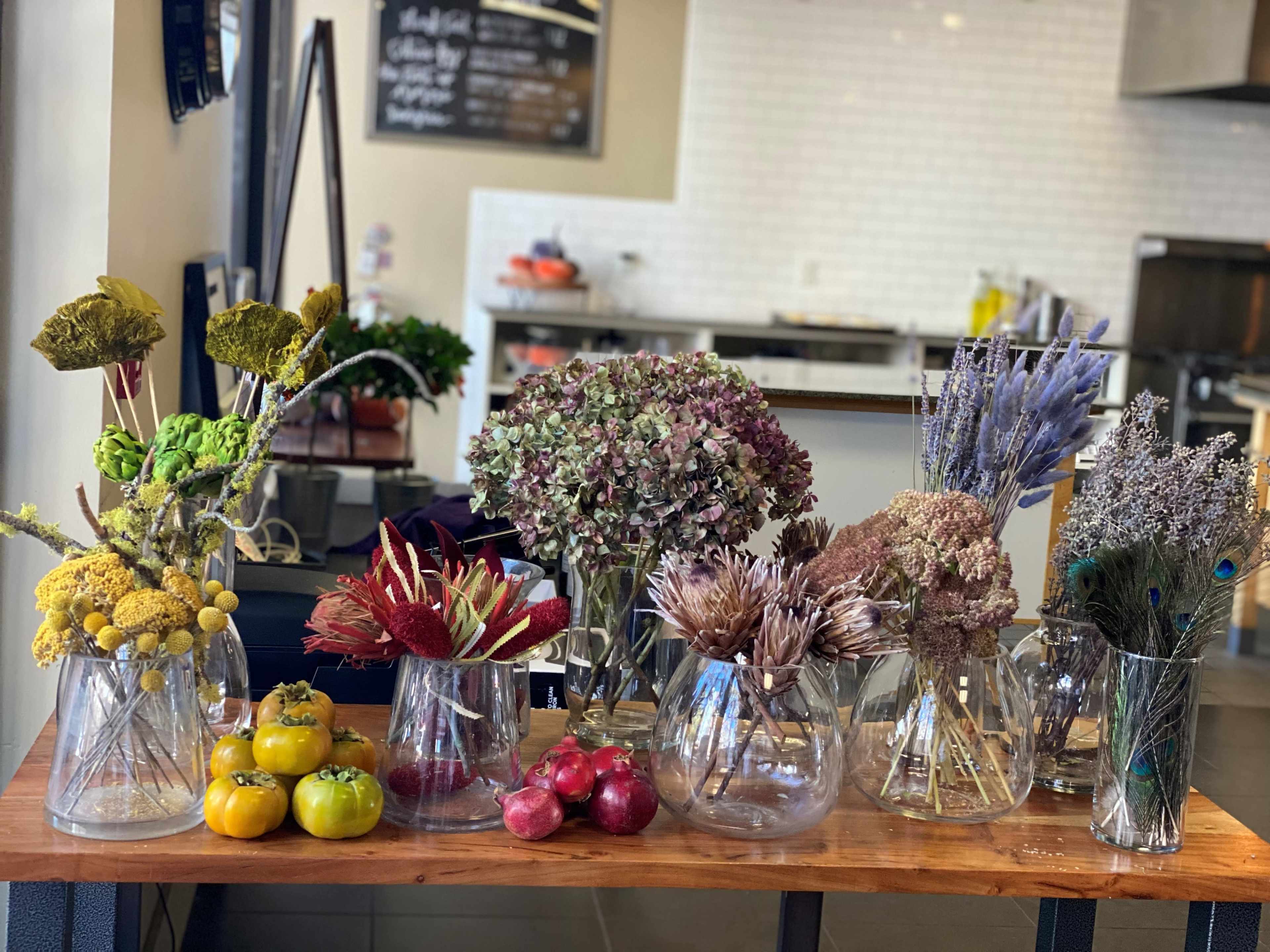 A wooden table displays various glass vases filled with dried flowers and fruits in a bright indoor setting.