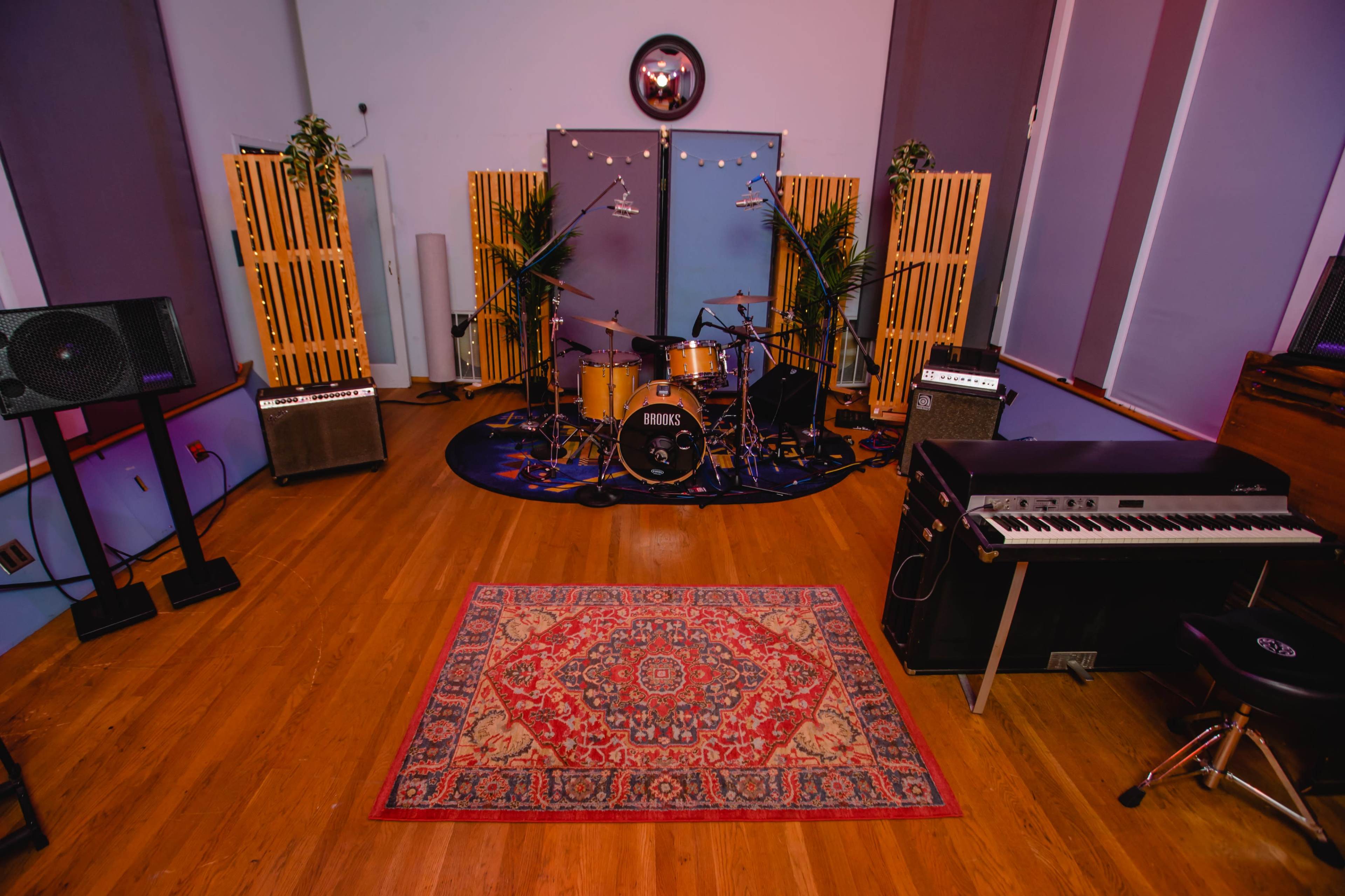 A music studio setup with a drum kit, amplifiers, and a keyboard on a patterned rug, surrounded by wooden acoustic panels.