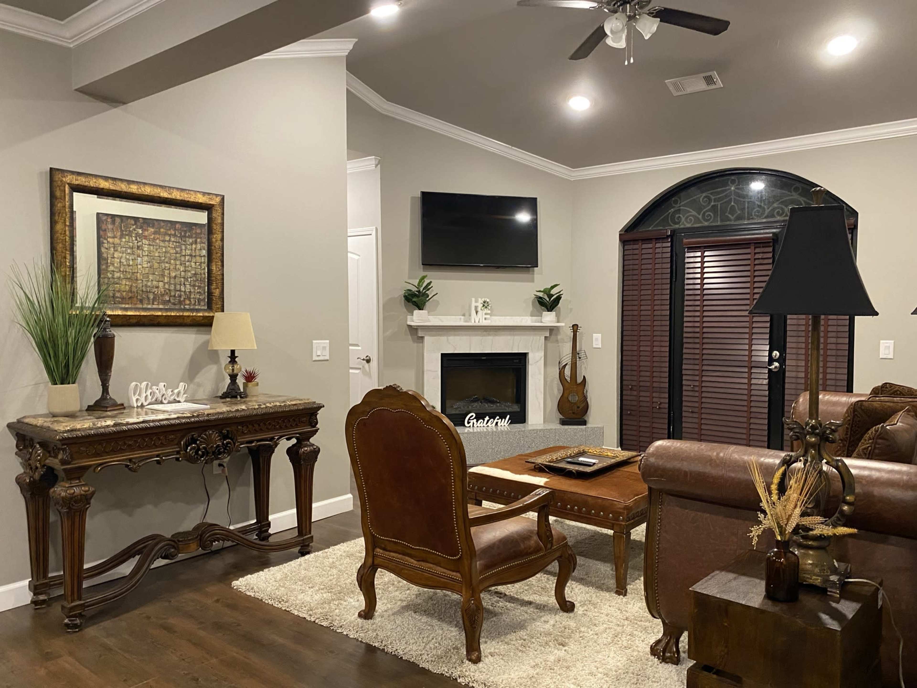 A spacious living room featuring a brown leather sofa, a wooden console table with decorative items, a fireplace with a television above it, and a chair positioned nearby.