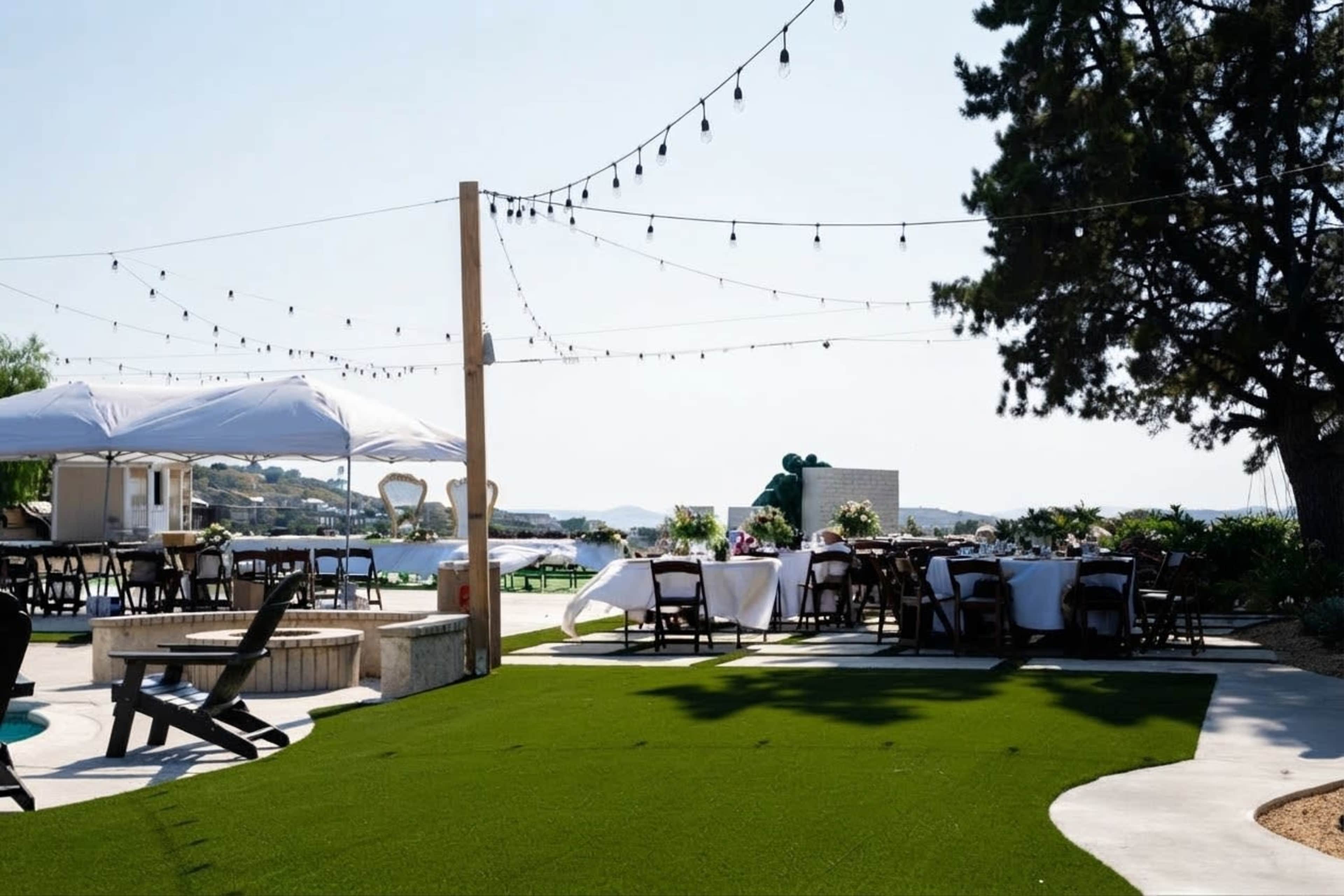 A patio area features tables set for an outdoor gathering, with string lights overhead and a view of a body of water in the background.
