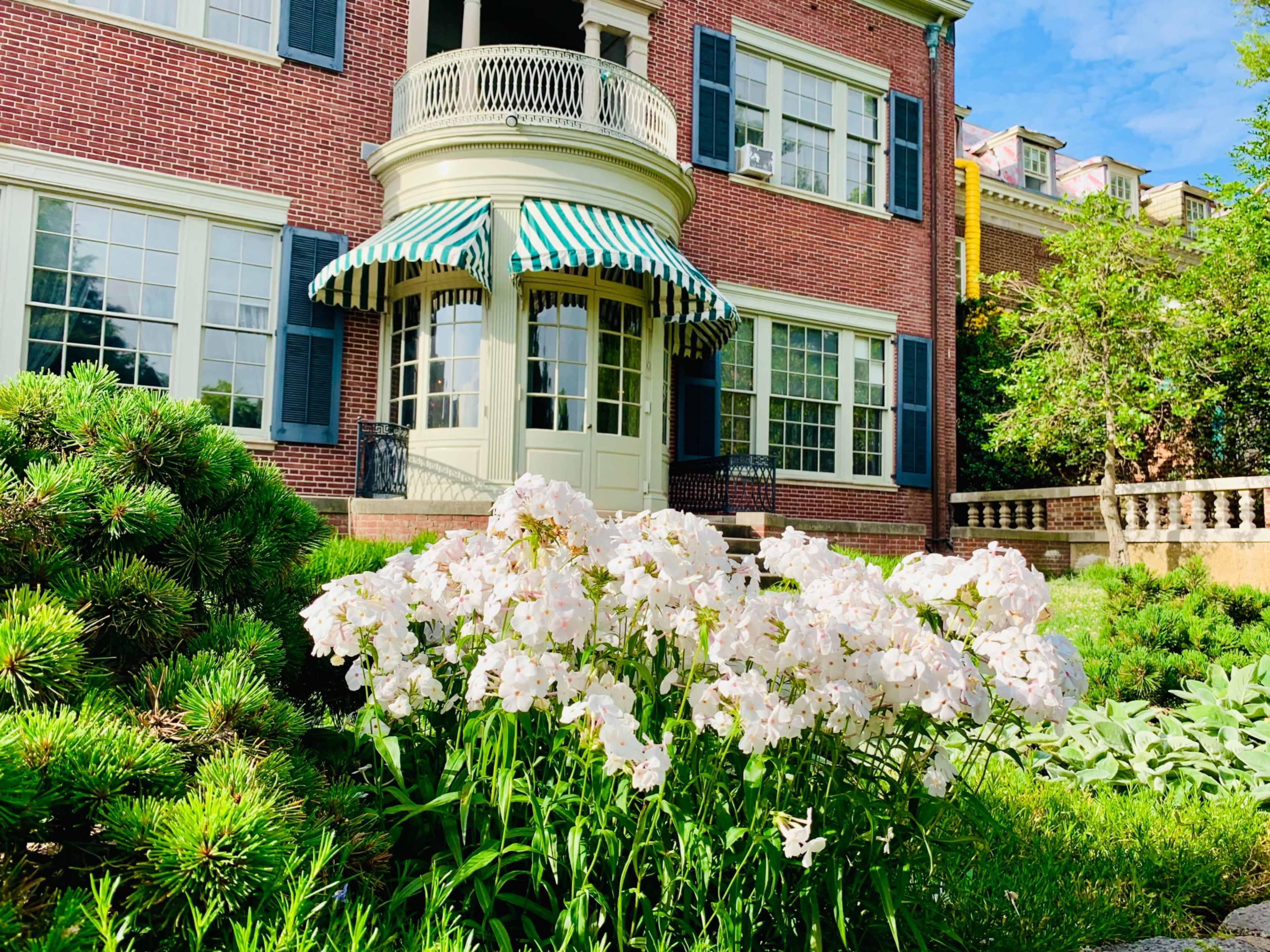 A brick house with green and white striped awnings features a curved porch, surrounded by a garden of white flowers and manicured shrubs.