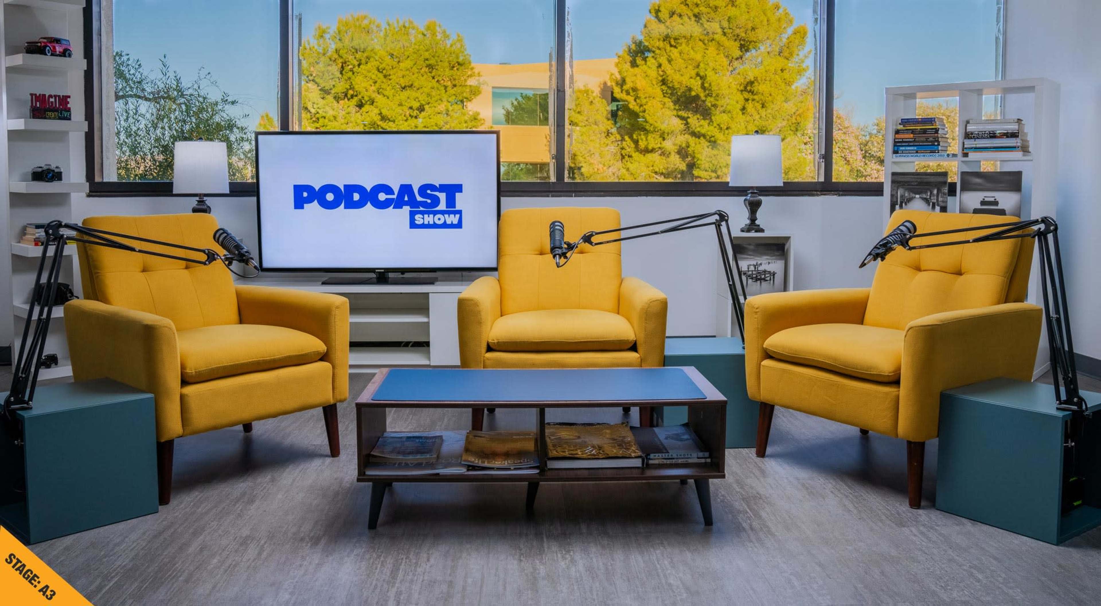 The image shows a well-lit podcast studio featuring two yellow chairs, a coffee table, and a television displaying the text "PODCAST SHOW," with microphones positioned in front of each chair.