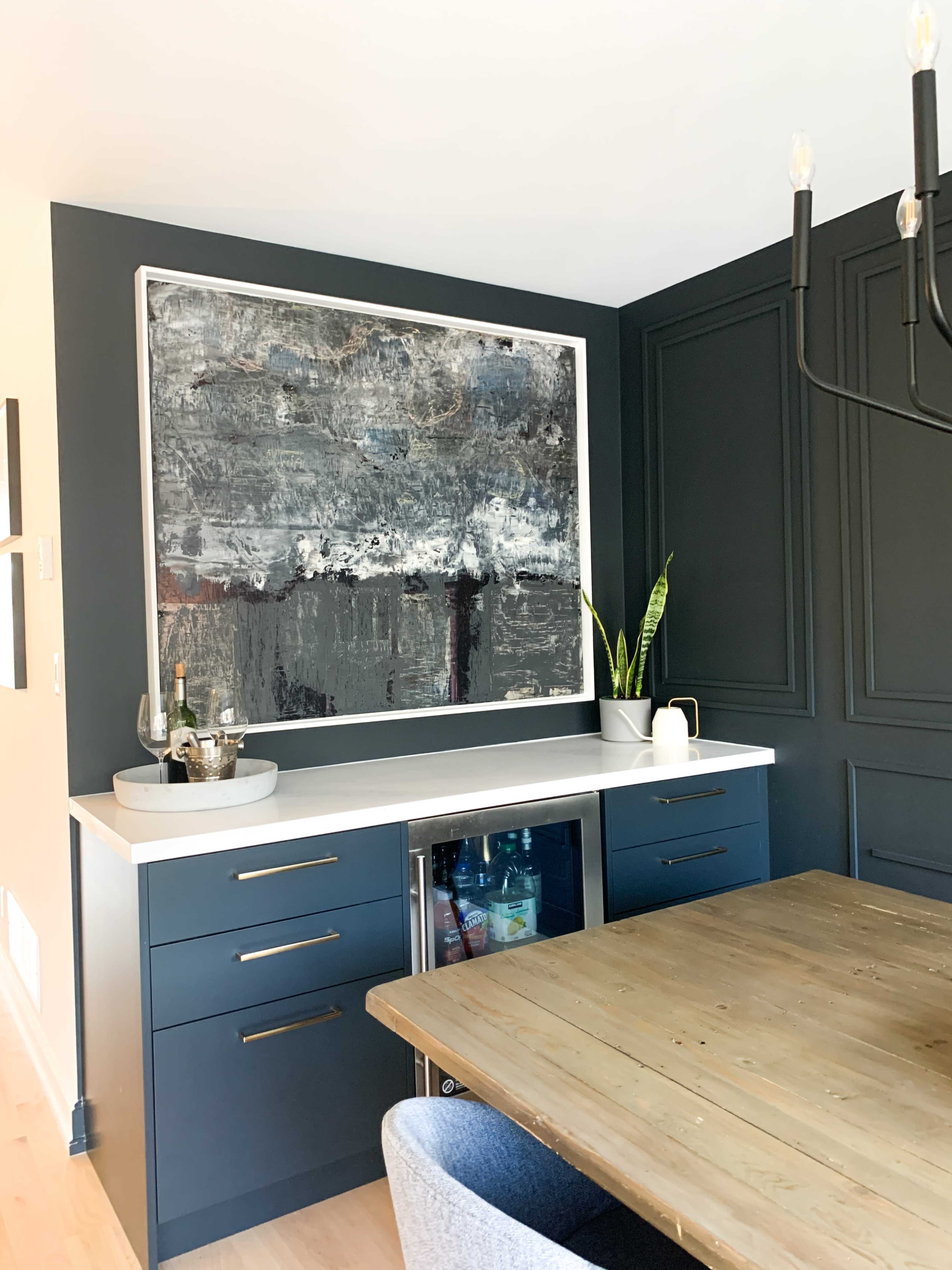 A modern dining area features a dark blue wall with a large abstract painting above a sideboard that includes a small fridge and a plant.