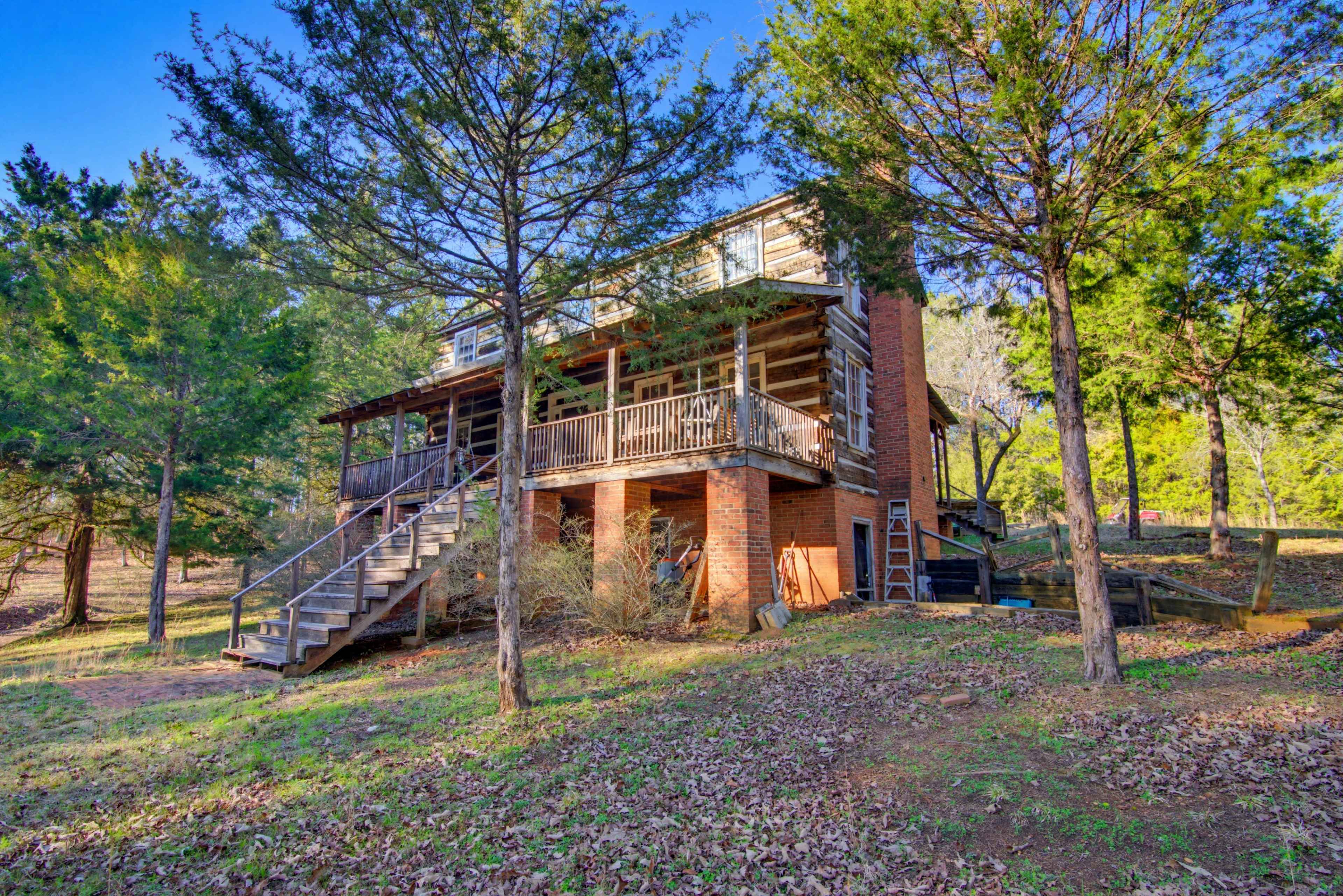 A two-story log cabin is situated on a wooded lot, with a staircase leading to a front porch.