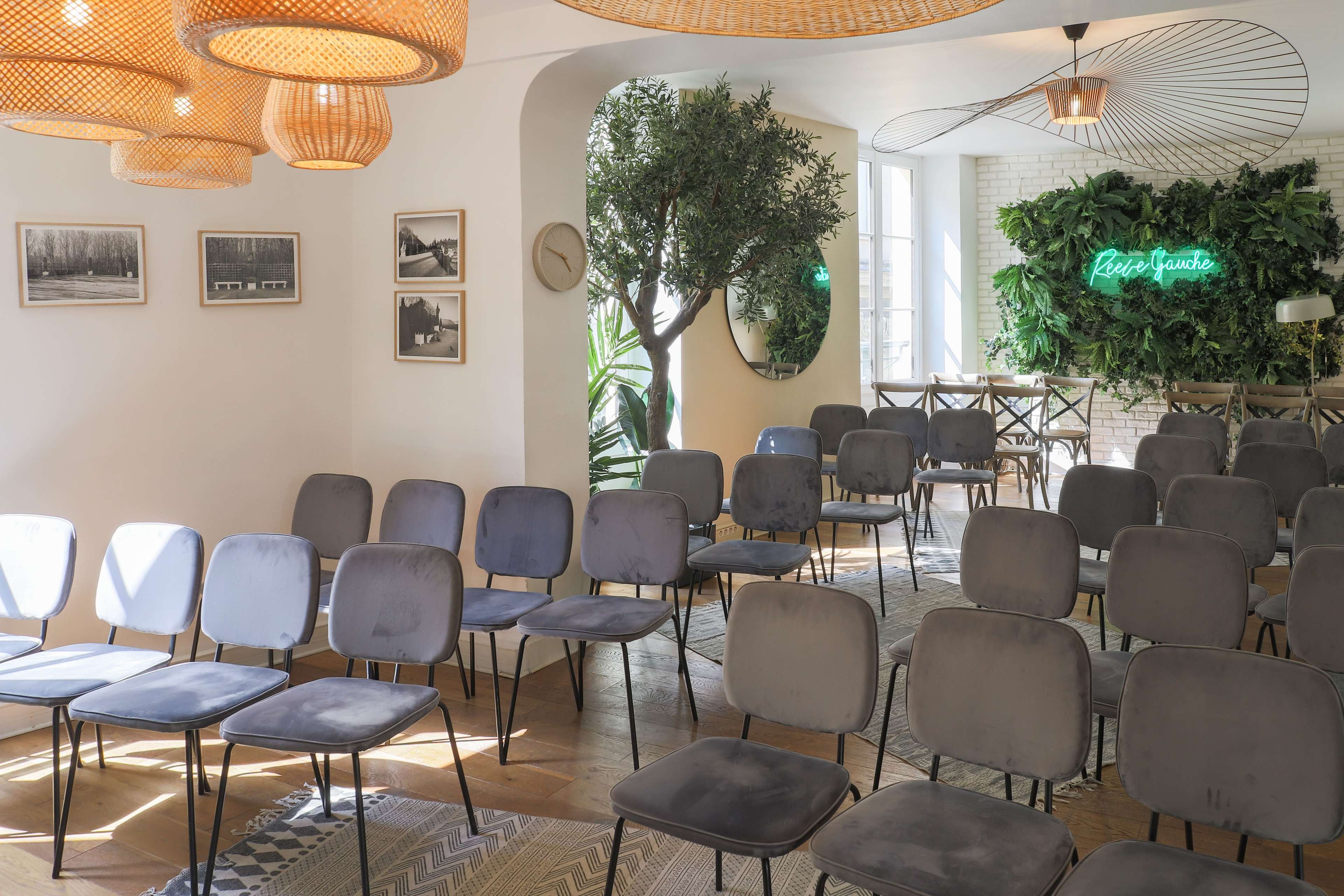 The image shows a spacious room arranged with rowed gray chairs, adorned with pendant lights and greenery along the walls.