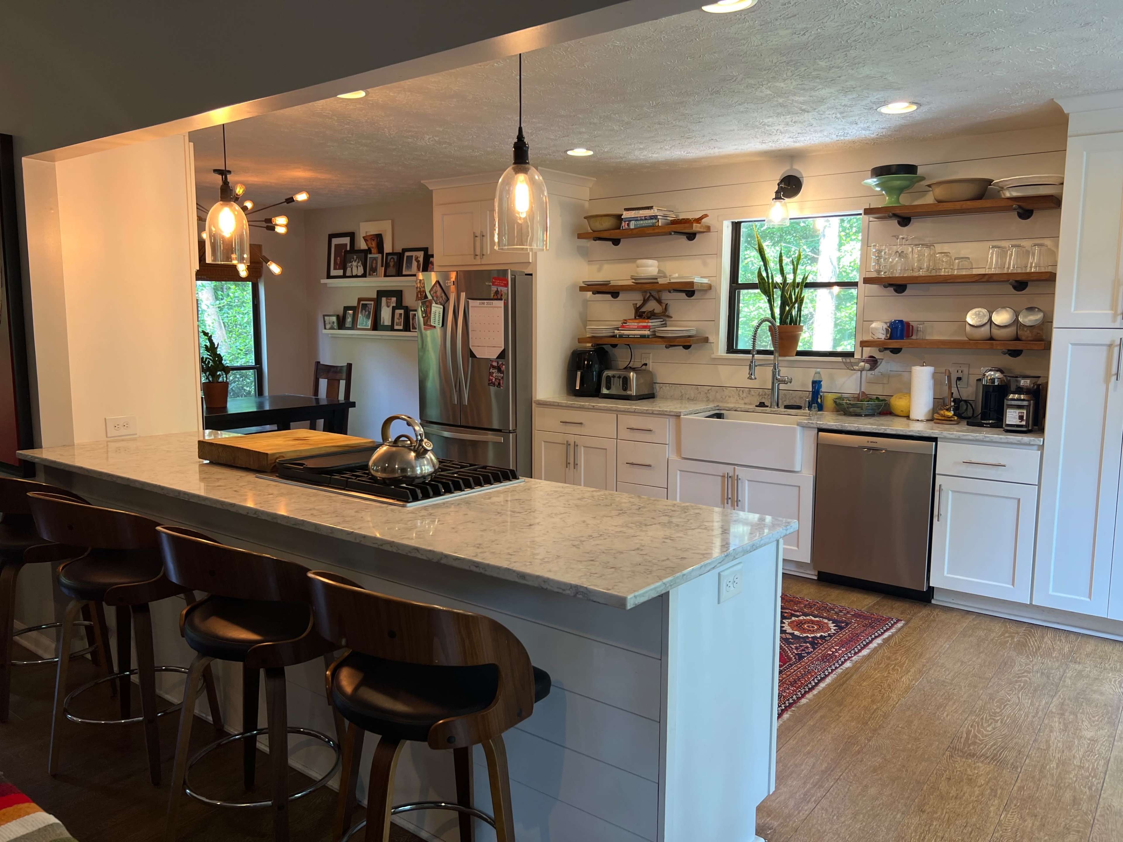 The image shows a modern kitchen with white cabinetry, an island with bar stools, and open shelving displaying various kitchen items.