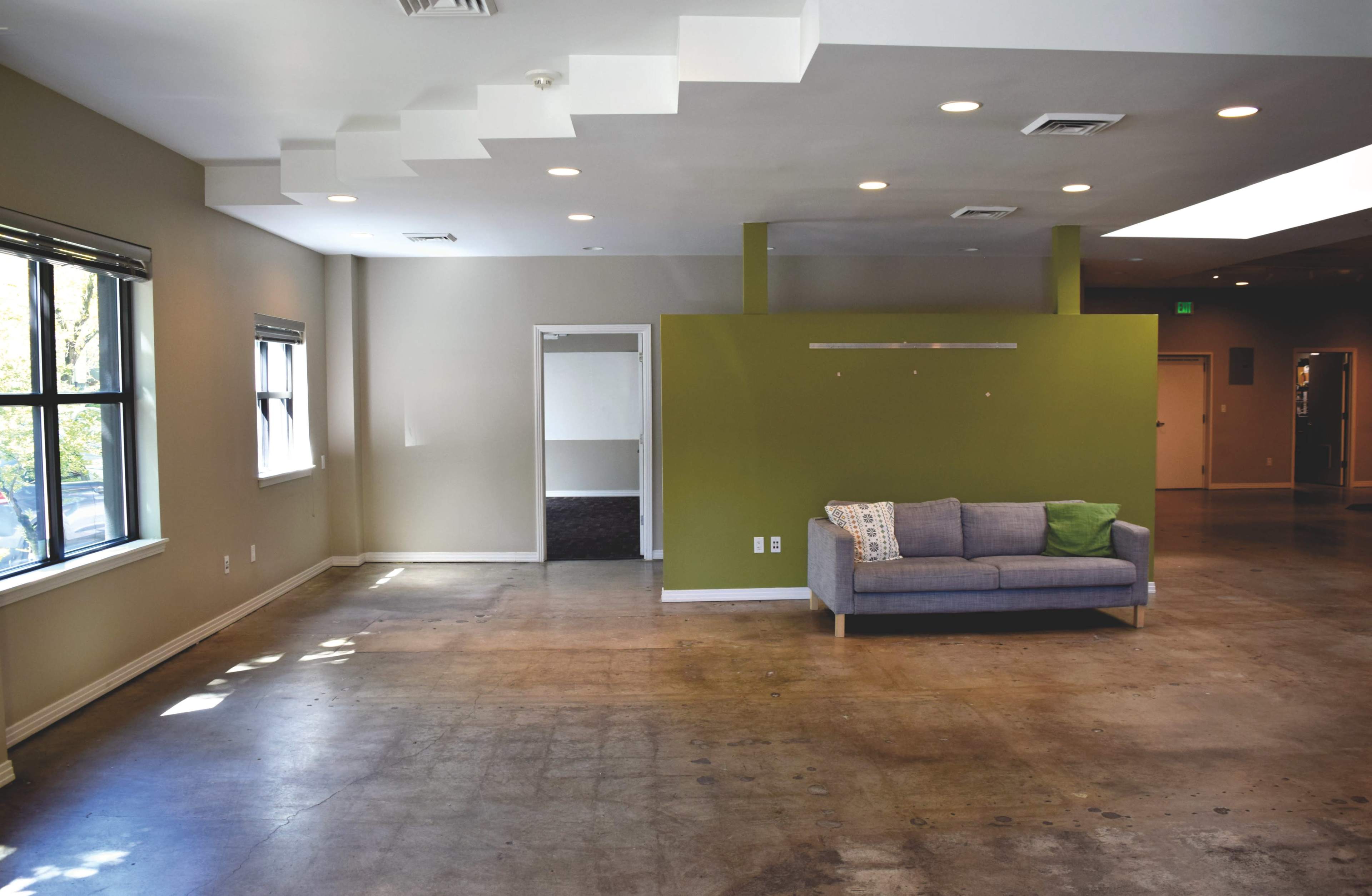 The image shows a spacious room with a concrete floor, a gray couch against a green accent wall, and a doorway leading into another area.