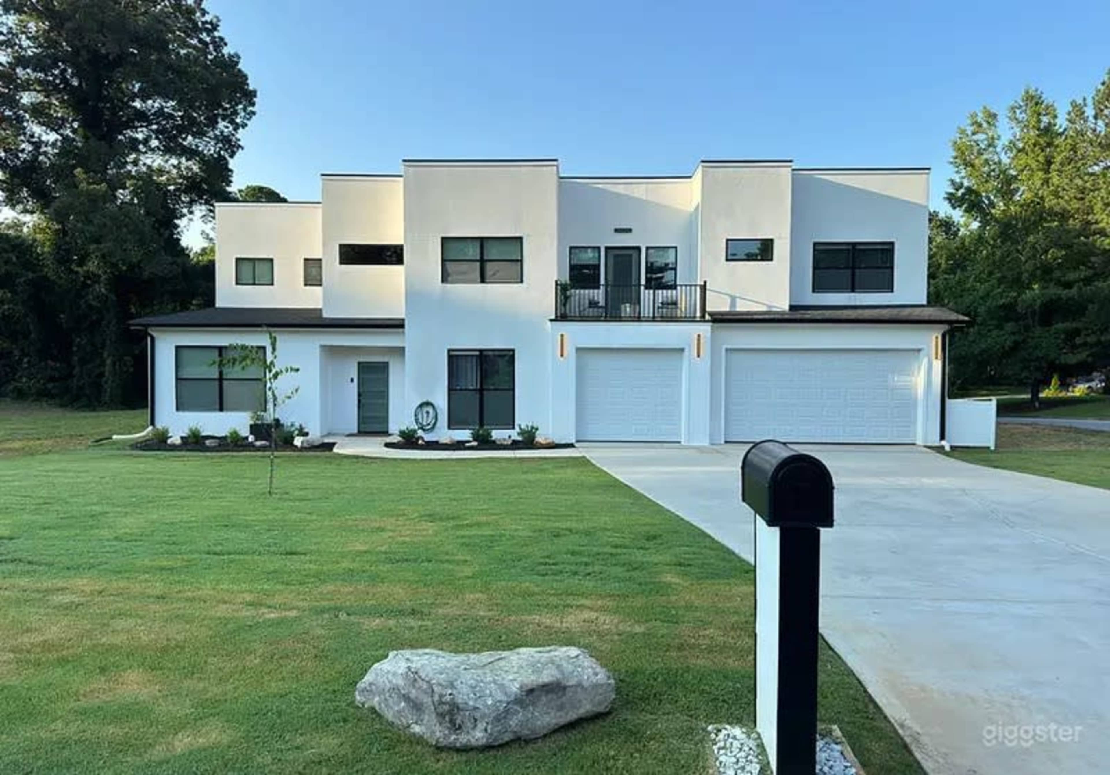 The image shows a modern, two-story white house with large windows and a neatly landscaped front yard, featuring a paved driveway and a mailbox in the foreground.