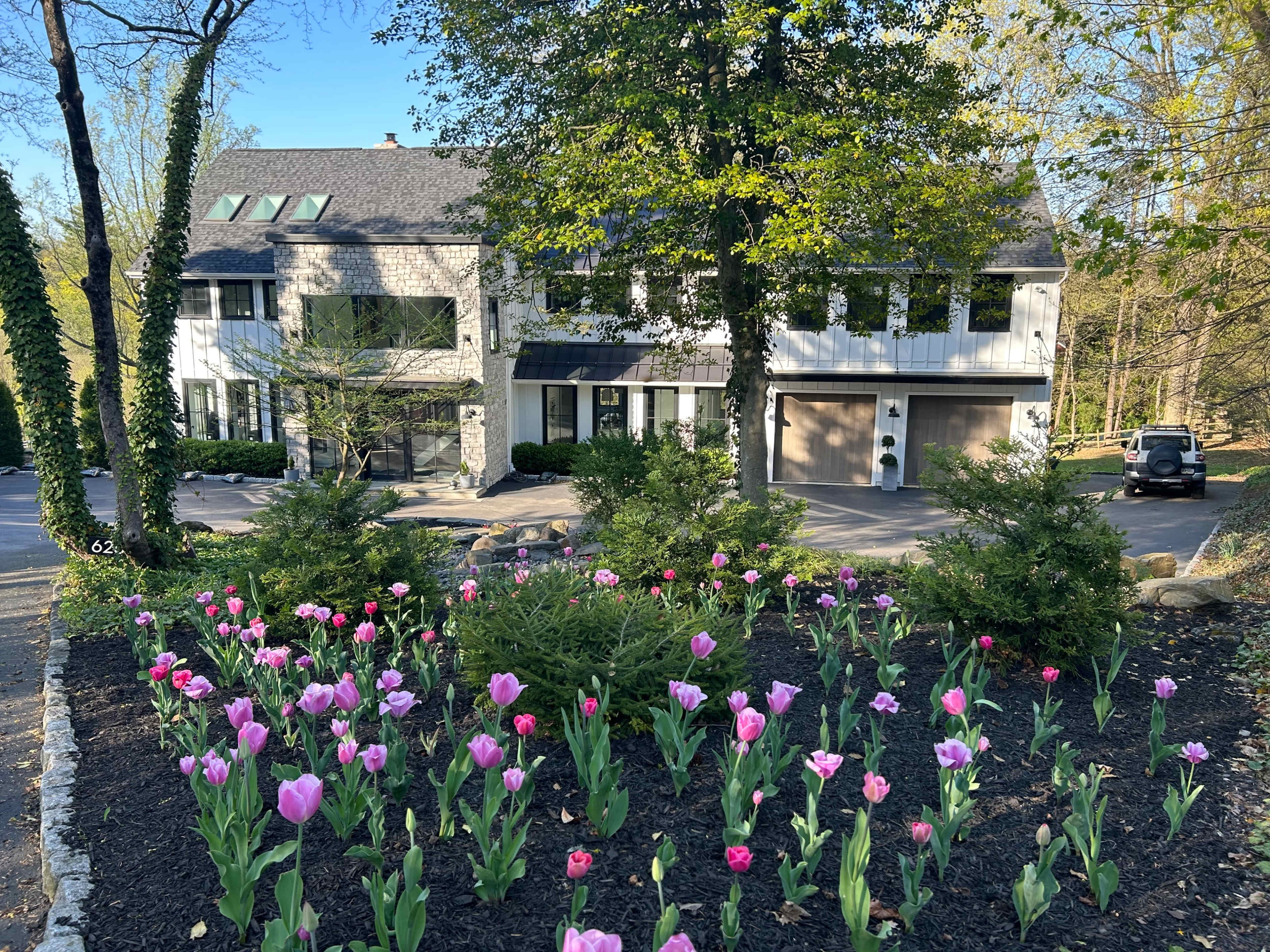 A house with a light exterior and large windows is set back from the street, surrounded by colorful tulip beds and trees.