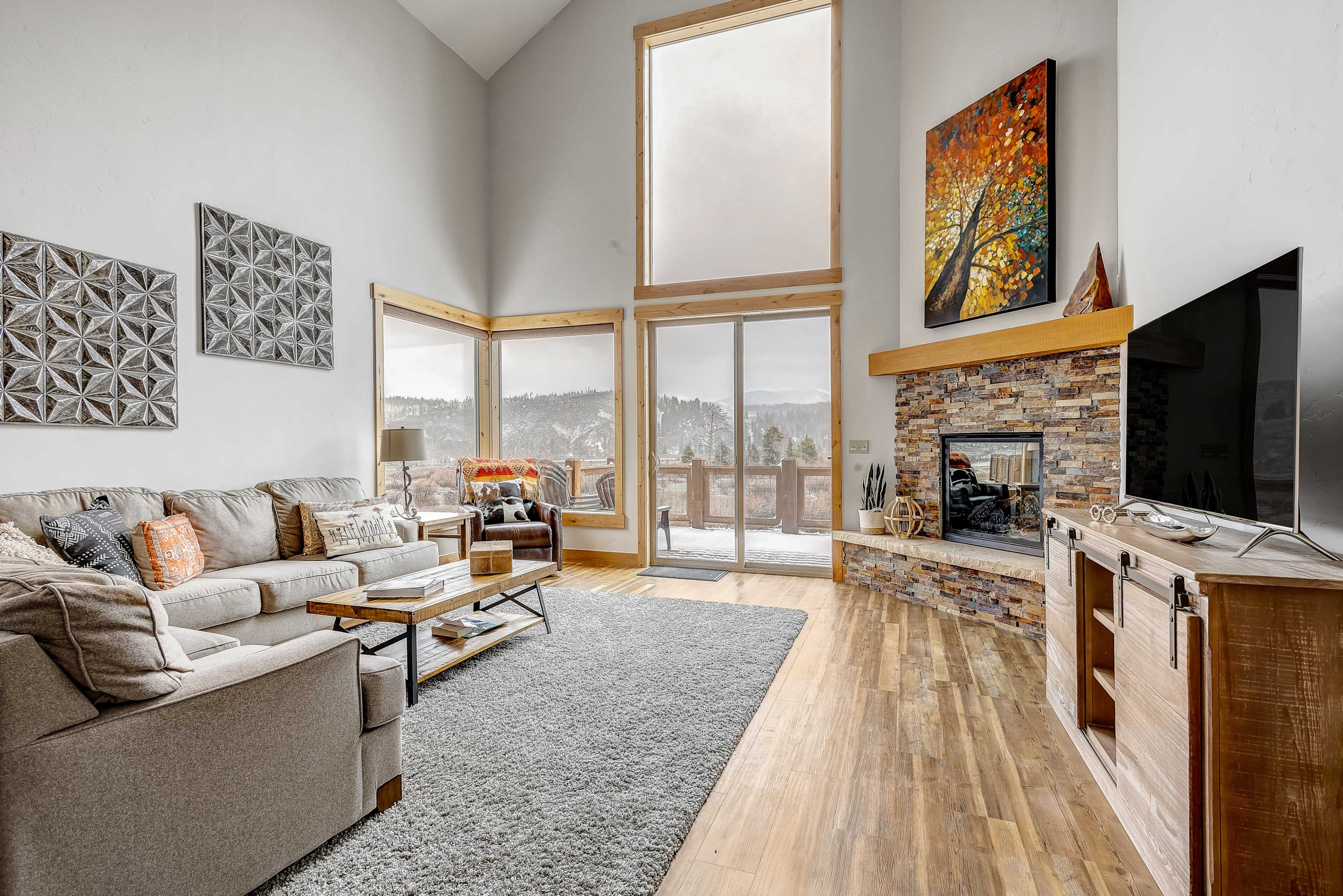 A spacious living room features a large window with a mountain view, a comfortable sectional sofa, a stone fireplace, and a television on a wooden cabinet.