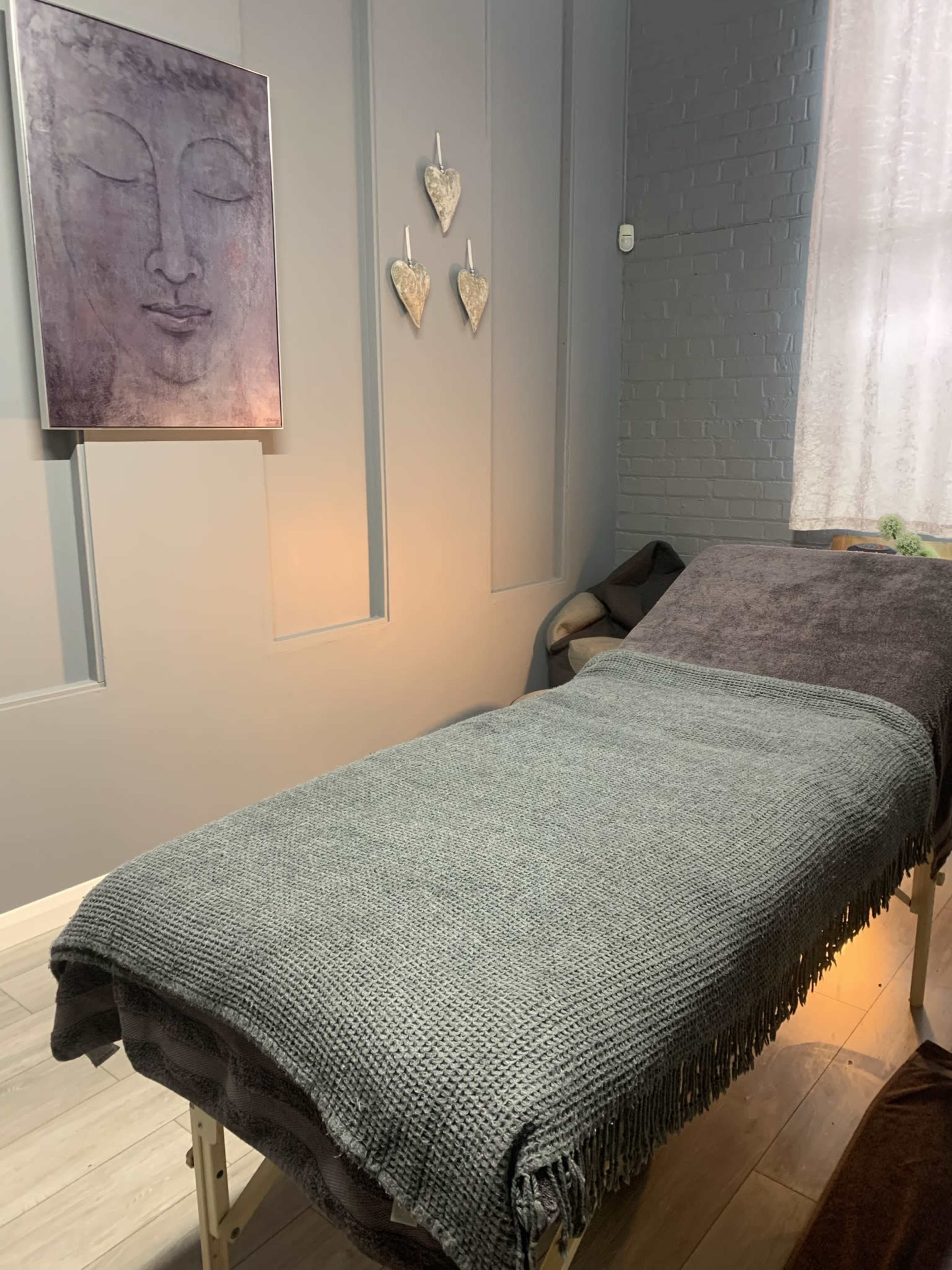 A wellness room with a massage table covered in a gray blanket, a framed artwork of a face on the wall, and decorative heart shapes hanging nearby.