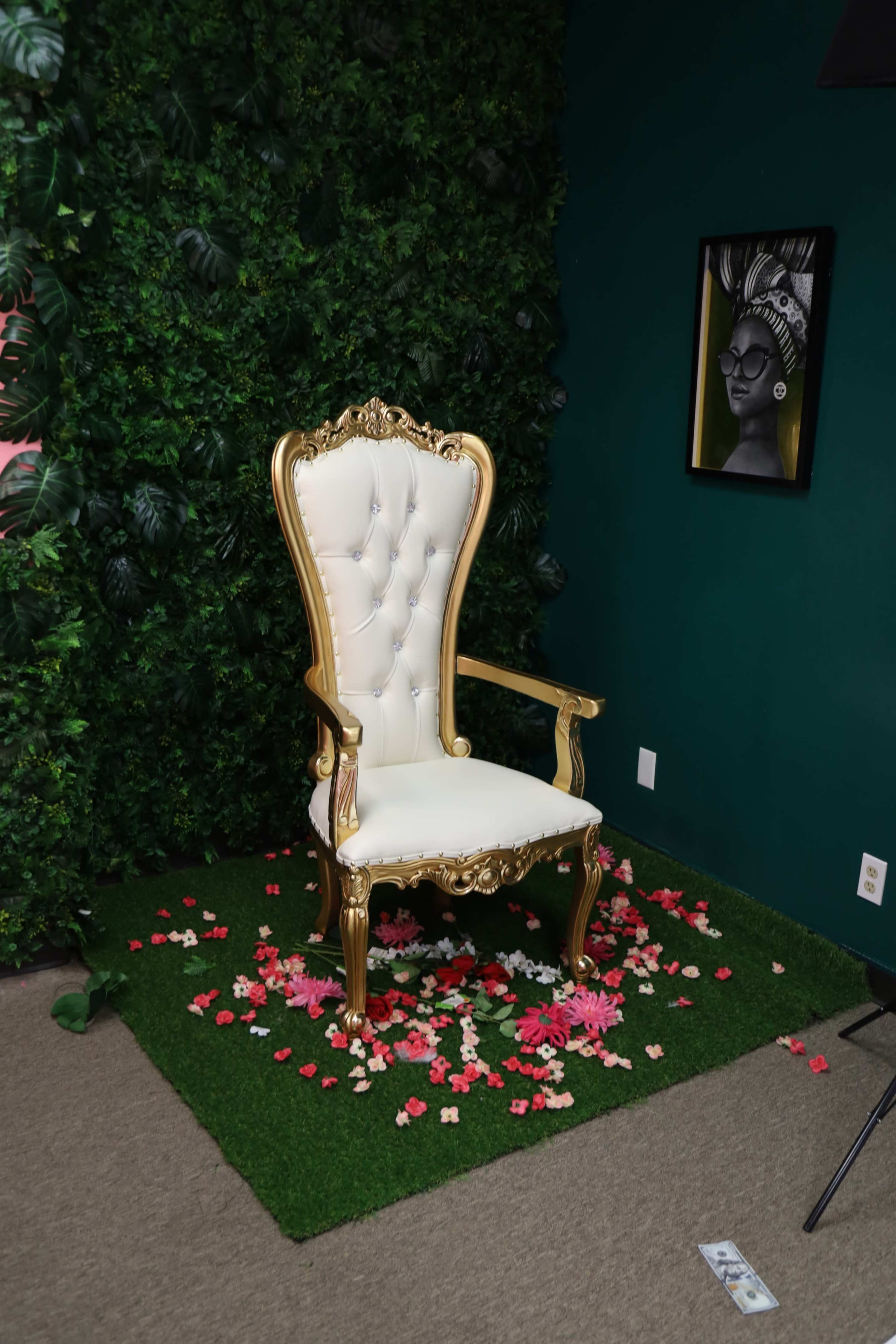 A white and gold chair is placed on a patch of grass surrounded by scattered pink and red flower petals against a green wall with greenery.