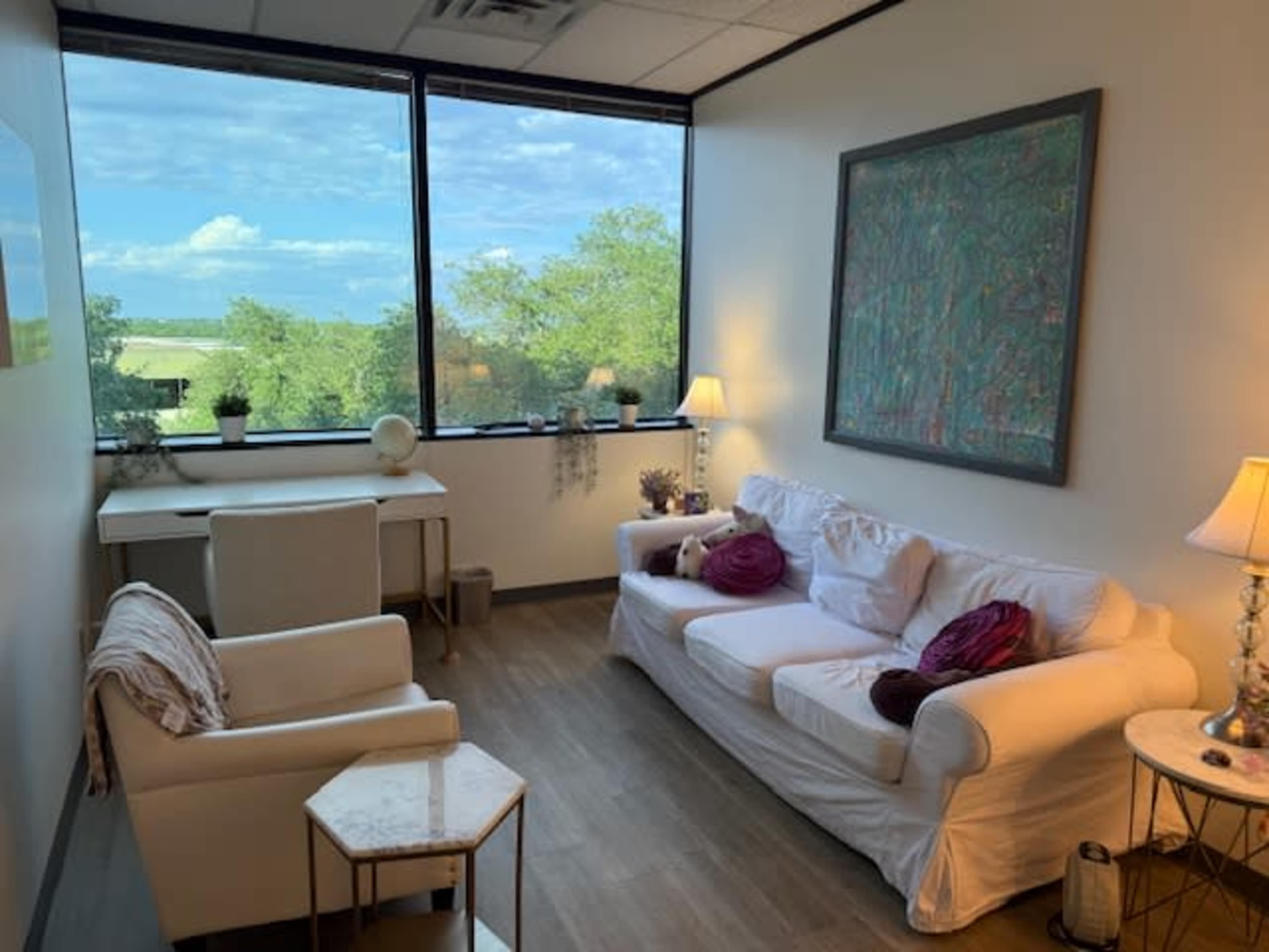 The image shows a small, well-lit room with a white couch, an armchair, a desk, and large windows offering a view of green trees outside.