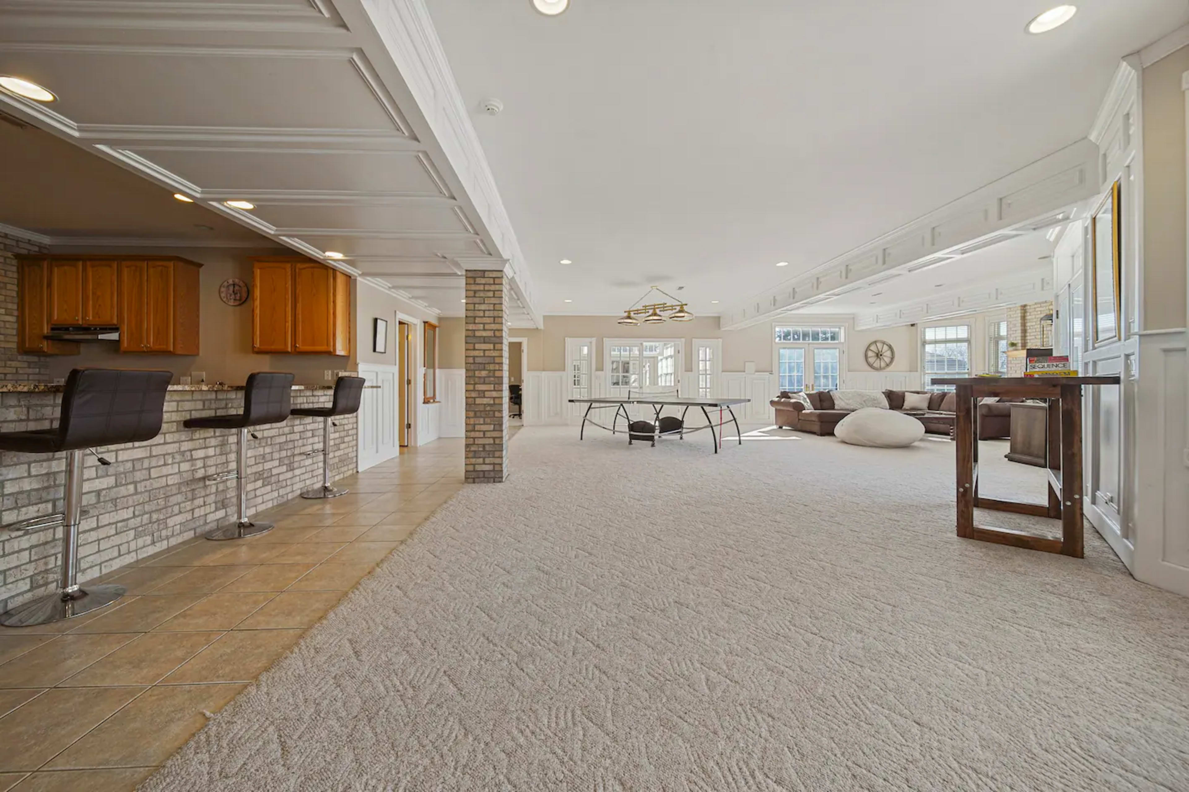 A spacious interior with a carpeted floor, featuring a bar area with stools on one side and a living area with large windows and a central table on the other.
