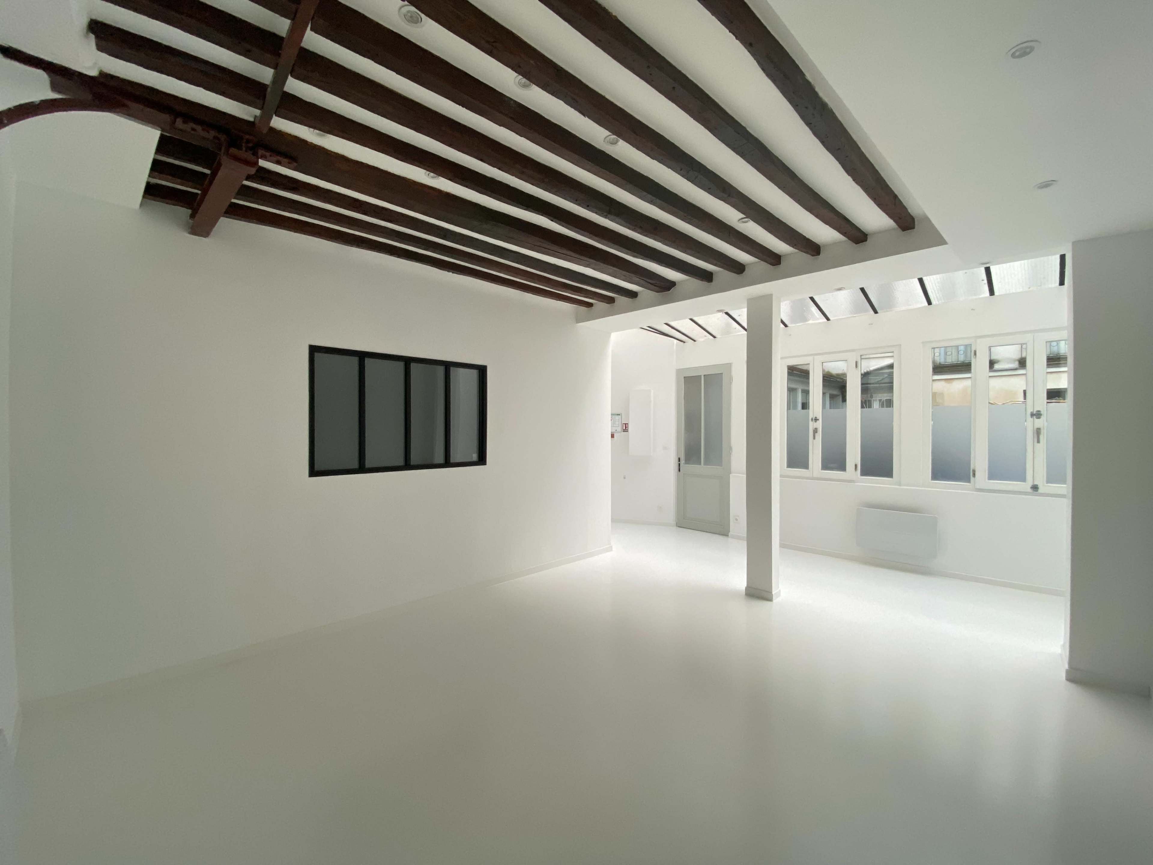 The image shows a bright, empty room with white walls, a white floor, exposed wooden beams on the ceiling, and large windows.