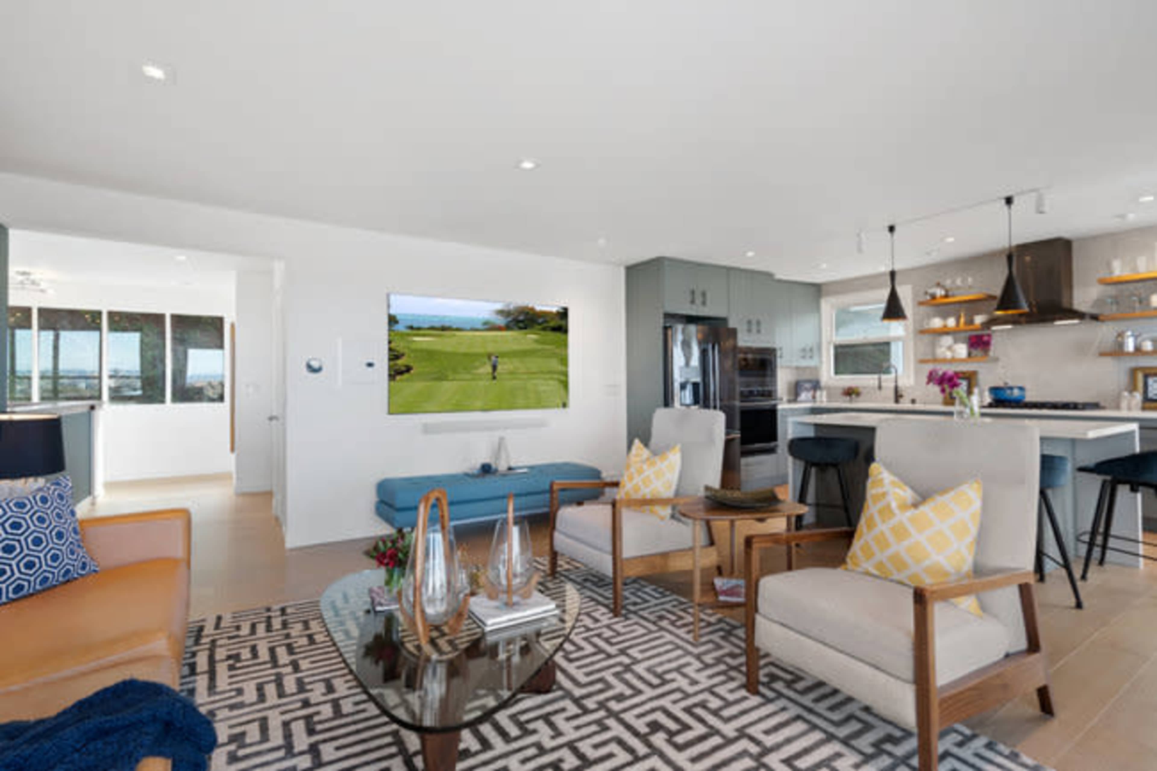 A modern living space with two armchairs, a glass coffee table, a wall-mounted television displaying a golf scene, and a kitchen area in the background.