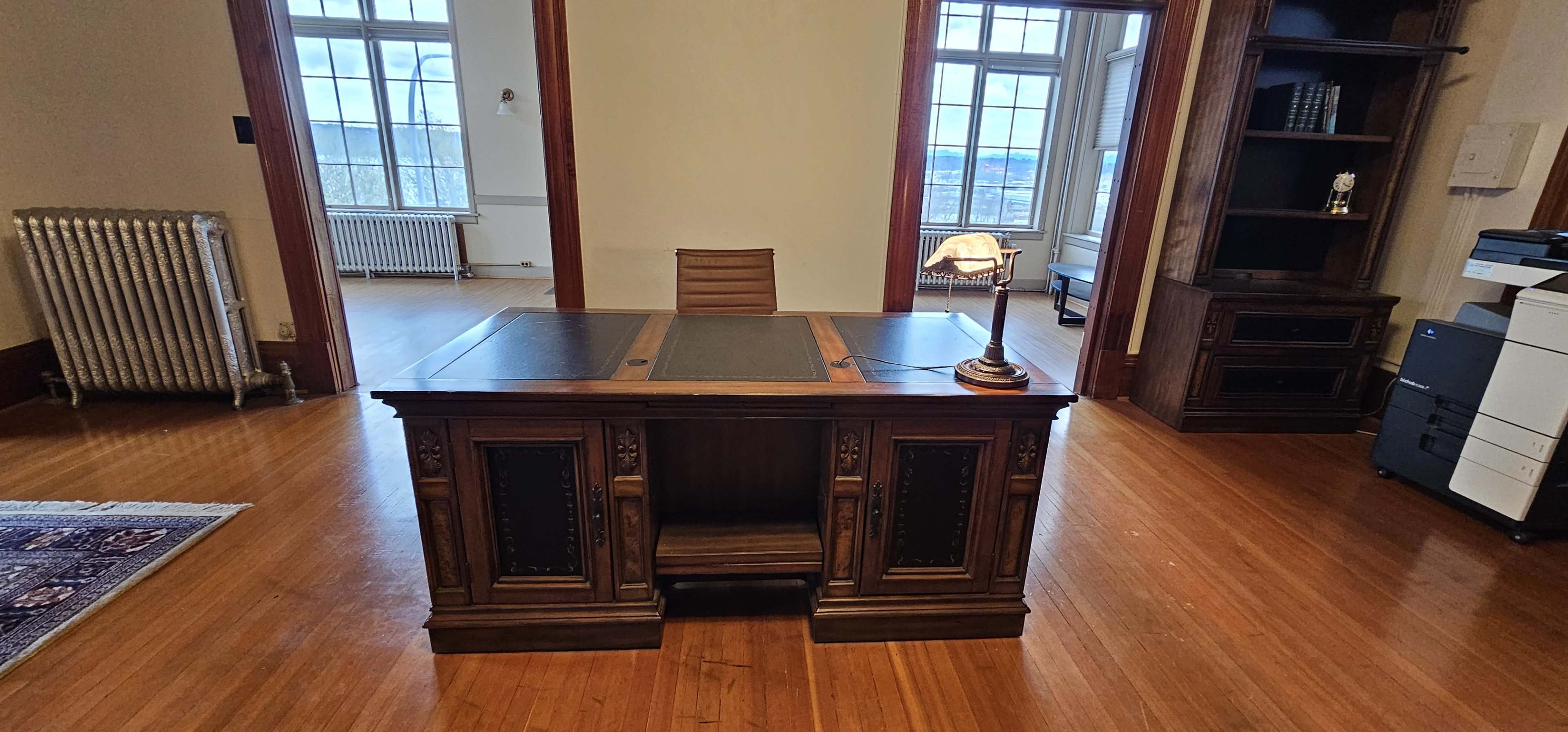 A wooden office desk with a lamp and chair in a room featuring large windows, a radiator, and a bookshelf.