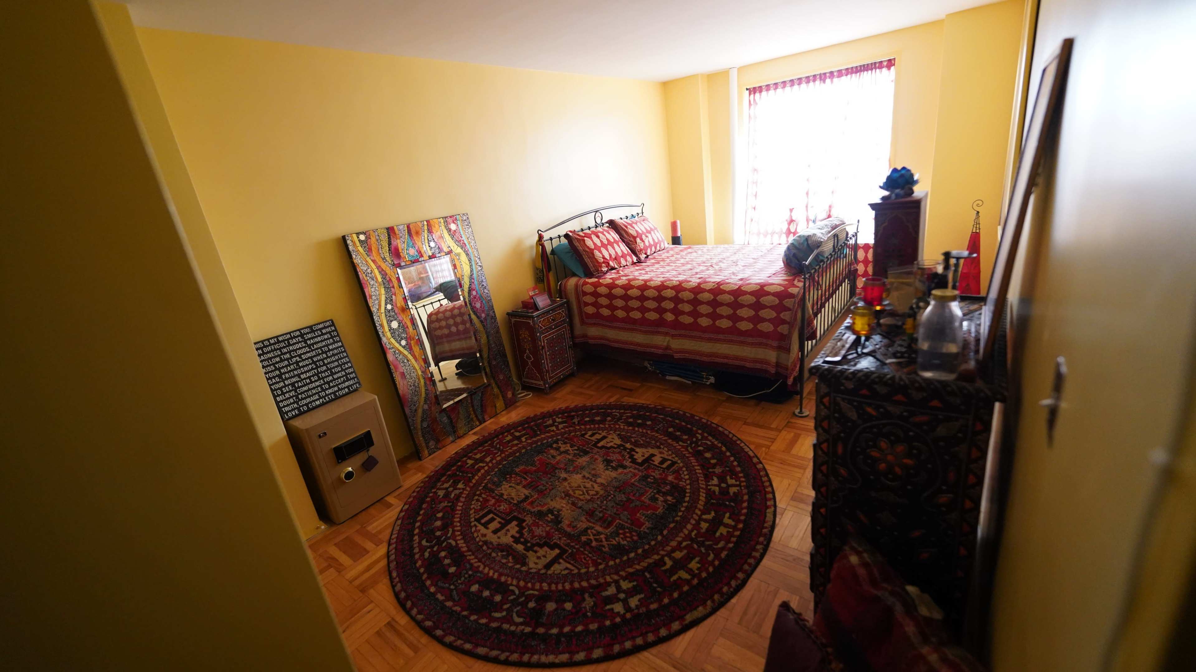 The image shows a cozy bedroom featuring a bed with patterned bedding, a large round rug on the floor, and a dresser beside a window with red curtains.