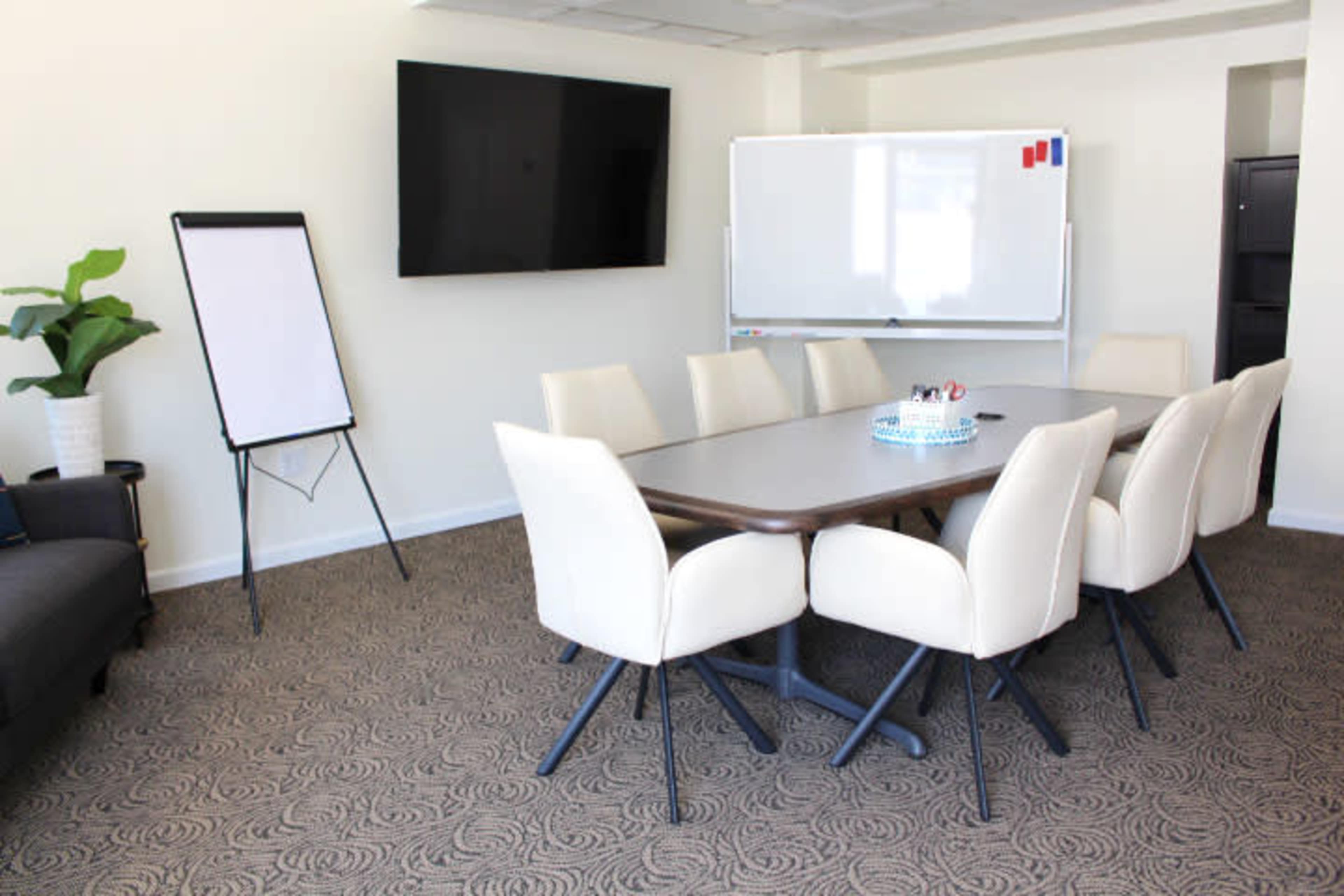 The image shows a modern conference room featuring a long table surrounded by white chairs, a large wall-mounted screen, a whiteboard, and a plant in the corner.