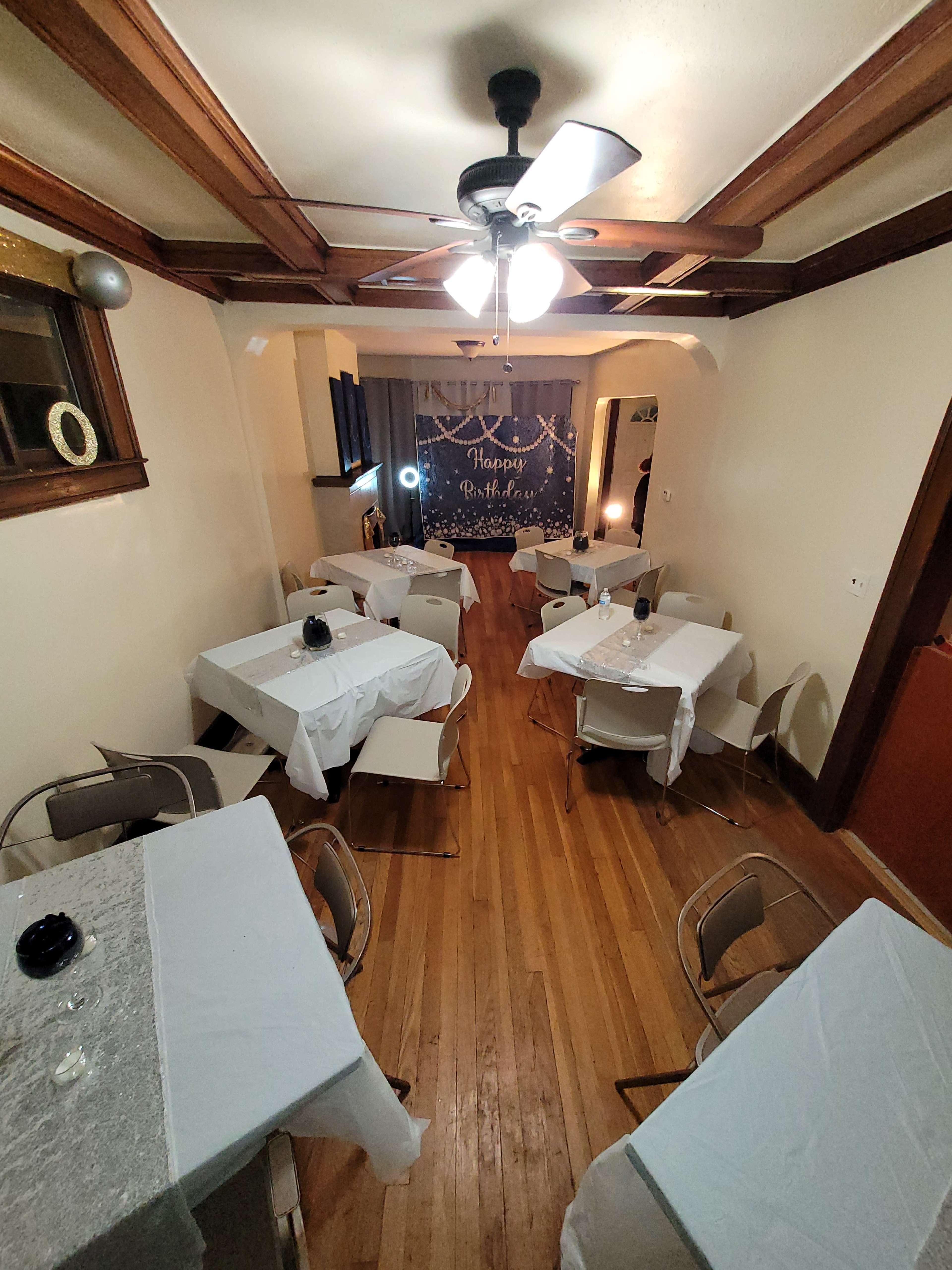 A dining area with several tables set for a birthday celebration, featuring white tablecloths and a decorative backdrop.