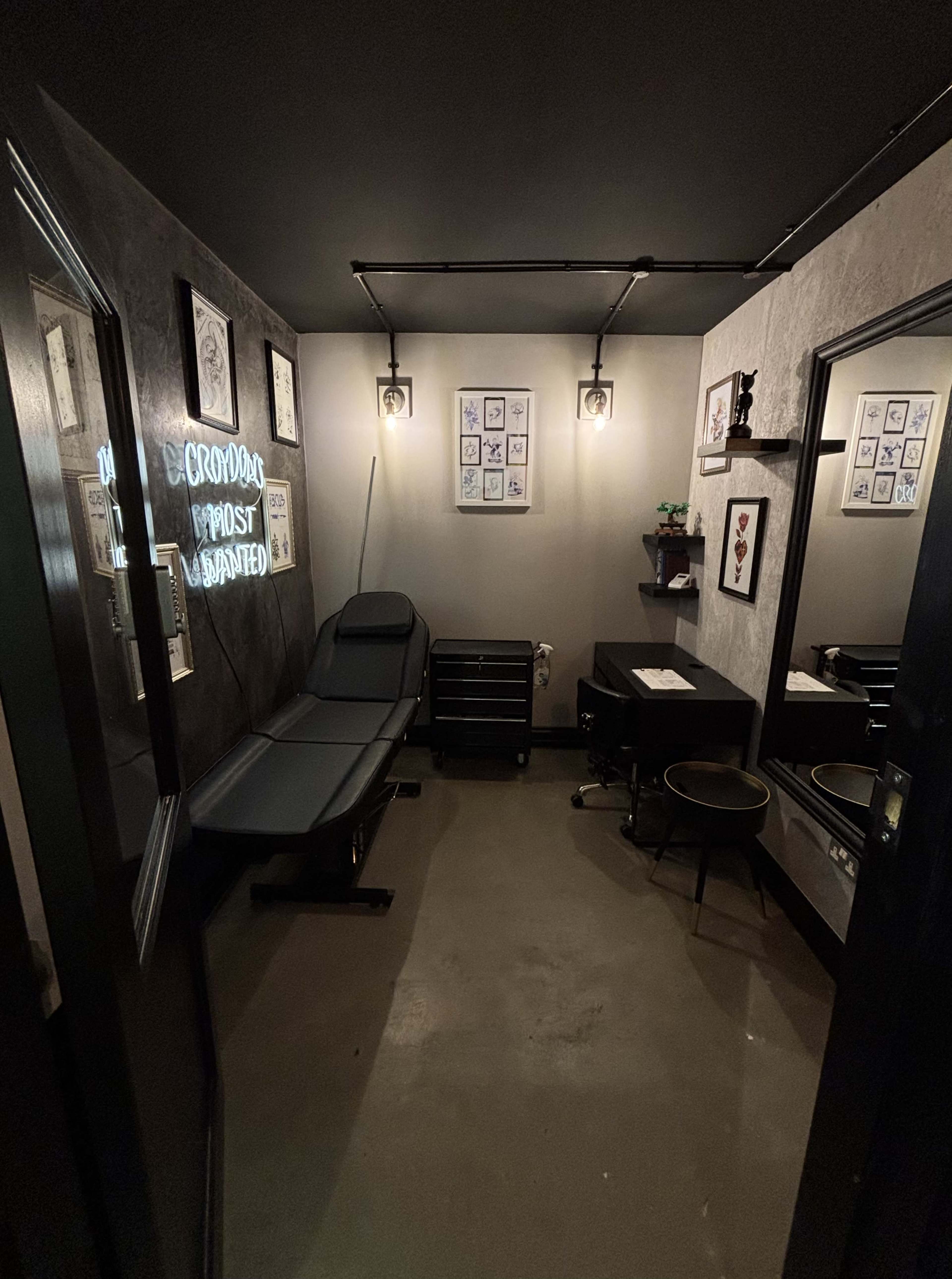 The image shows a small tattoo studio with a reclining chair, a work table, and framed tattoo designs on the walls.